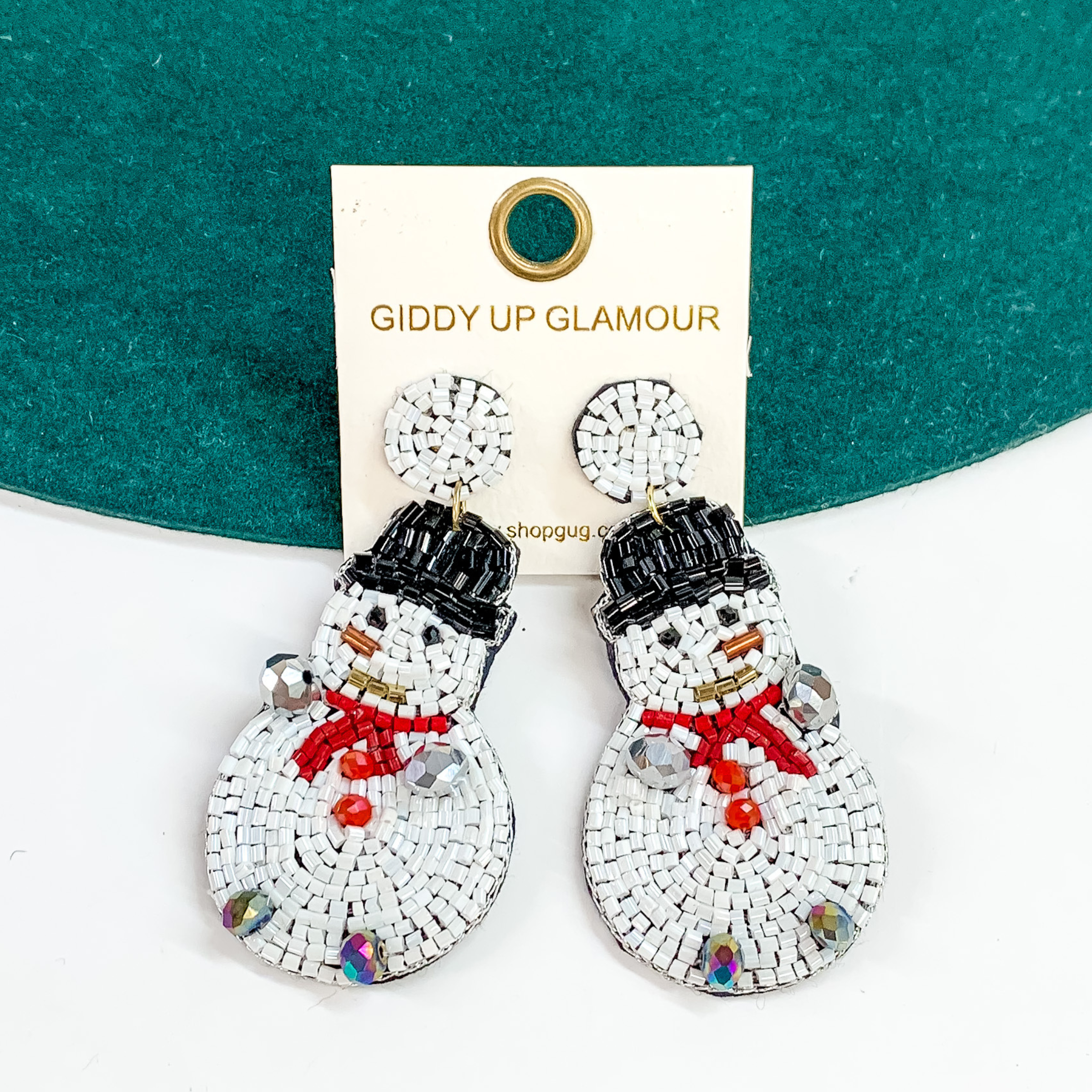 White beaded dangle snowman earrings. These earrings include the face of the snowman, a top hat, and beads for hands and feet. These earrings are pictured partially laying on a green hat brim on a white background. 