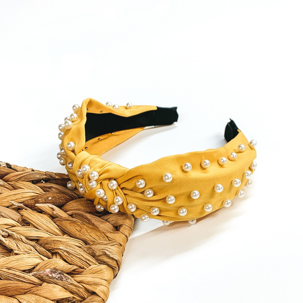 Yellow headband with white pearl accents. This headband is pictured laying partially on a basket weave board on a white background.