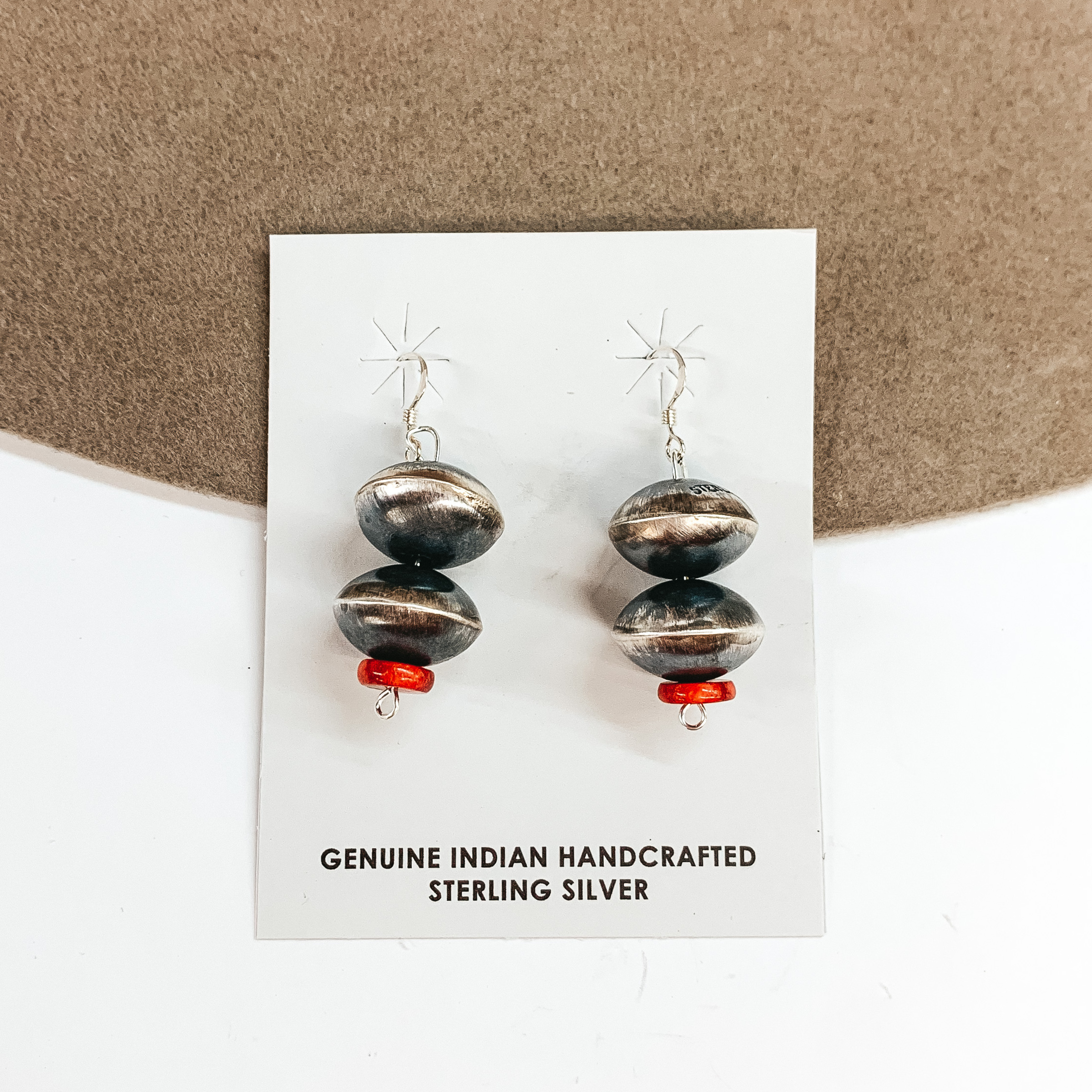 The earrings pictured include fish hook earrings with a two saucer pearl drop and a red stone at the bottom. These earrings are pictured on a tan and white background. 