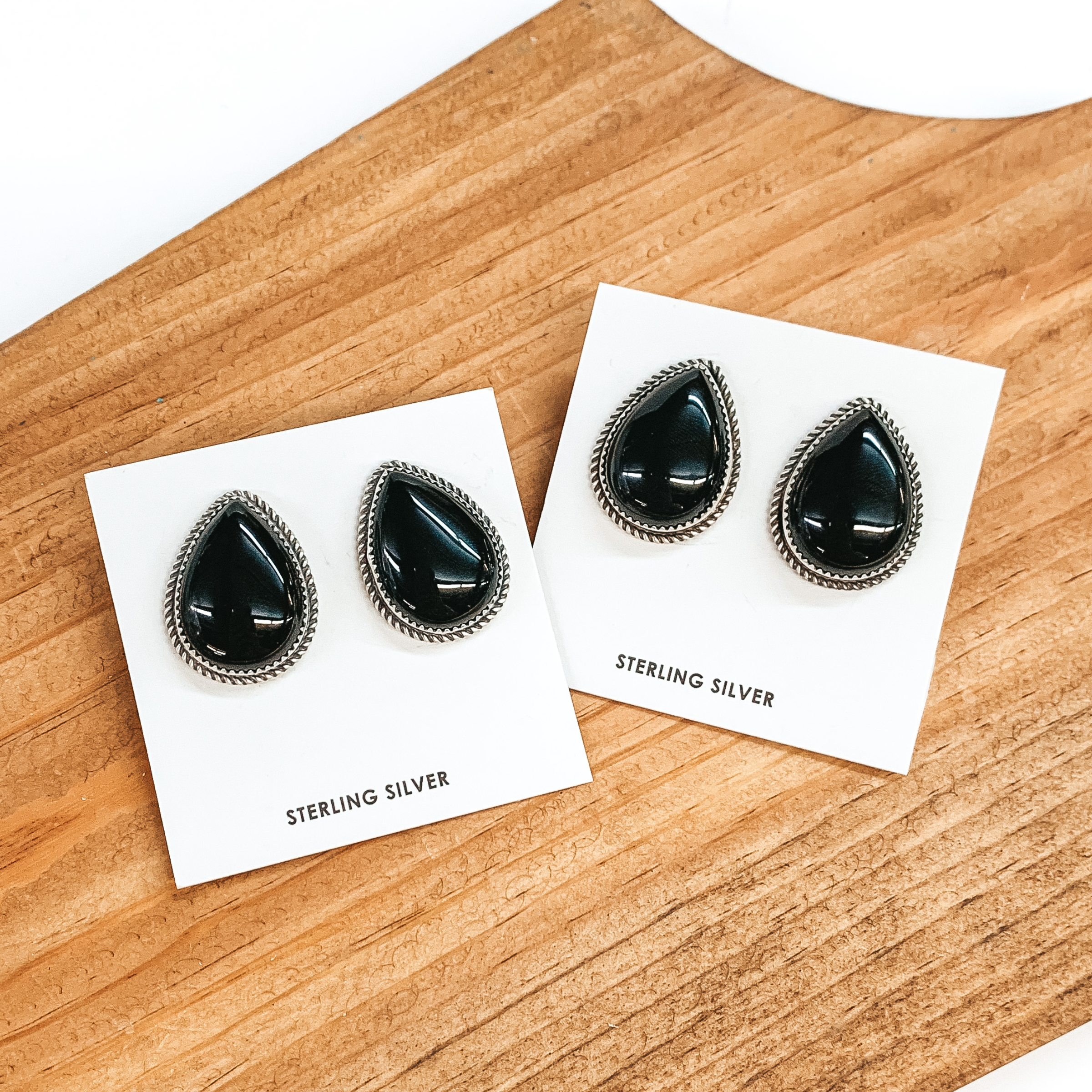 Two pairs of teardrop stud earrings that include a silver backing and black onyx stones. These earrings are pictured on a brown block on a white background. 