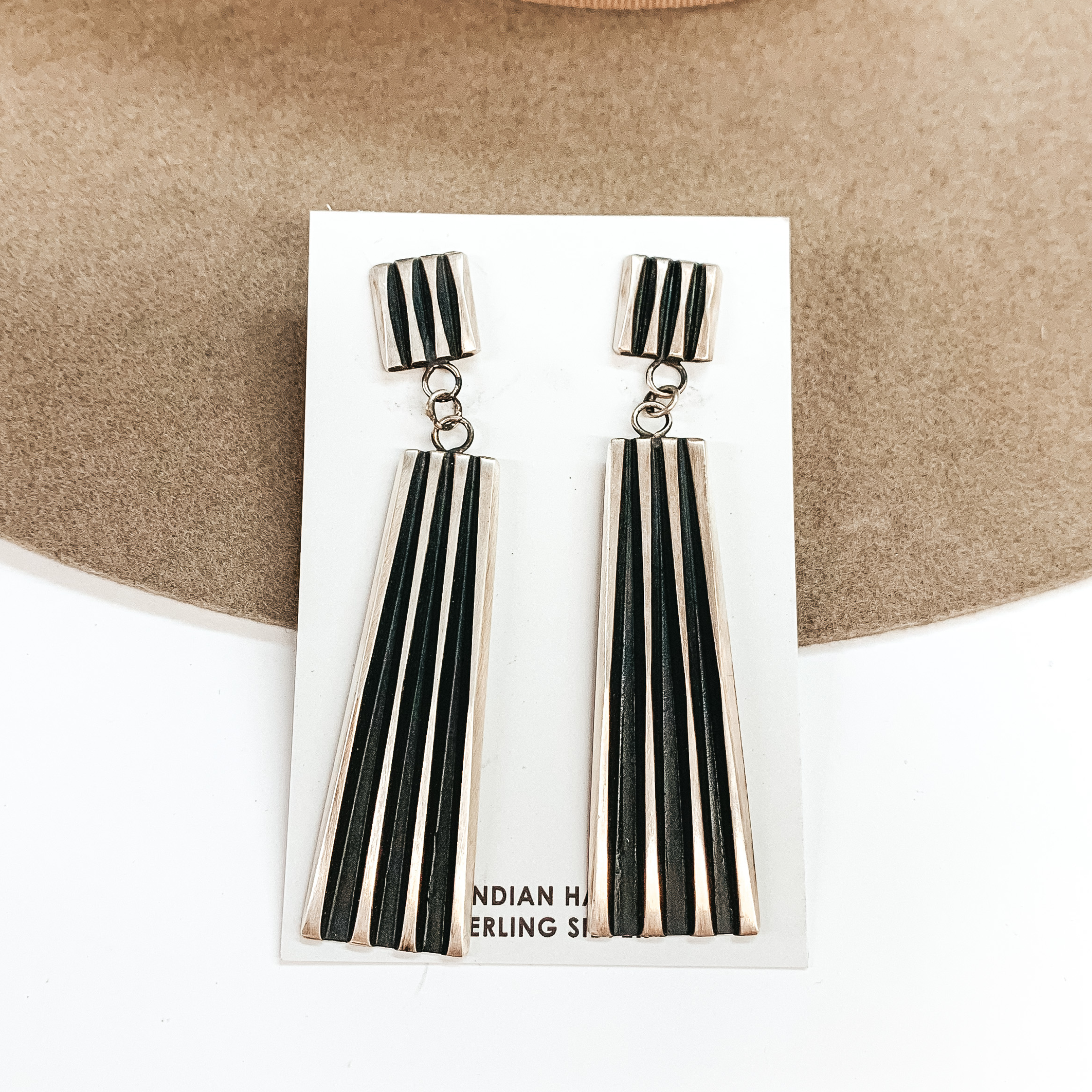 Sterling silver post back earrings with a drop and include some cutout lines. These earrings are pictured on a tan and white background. 