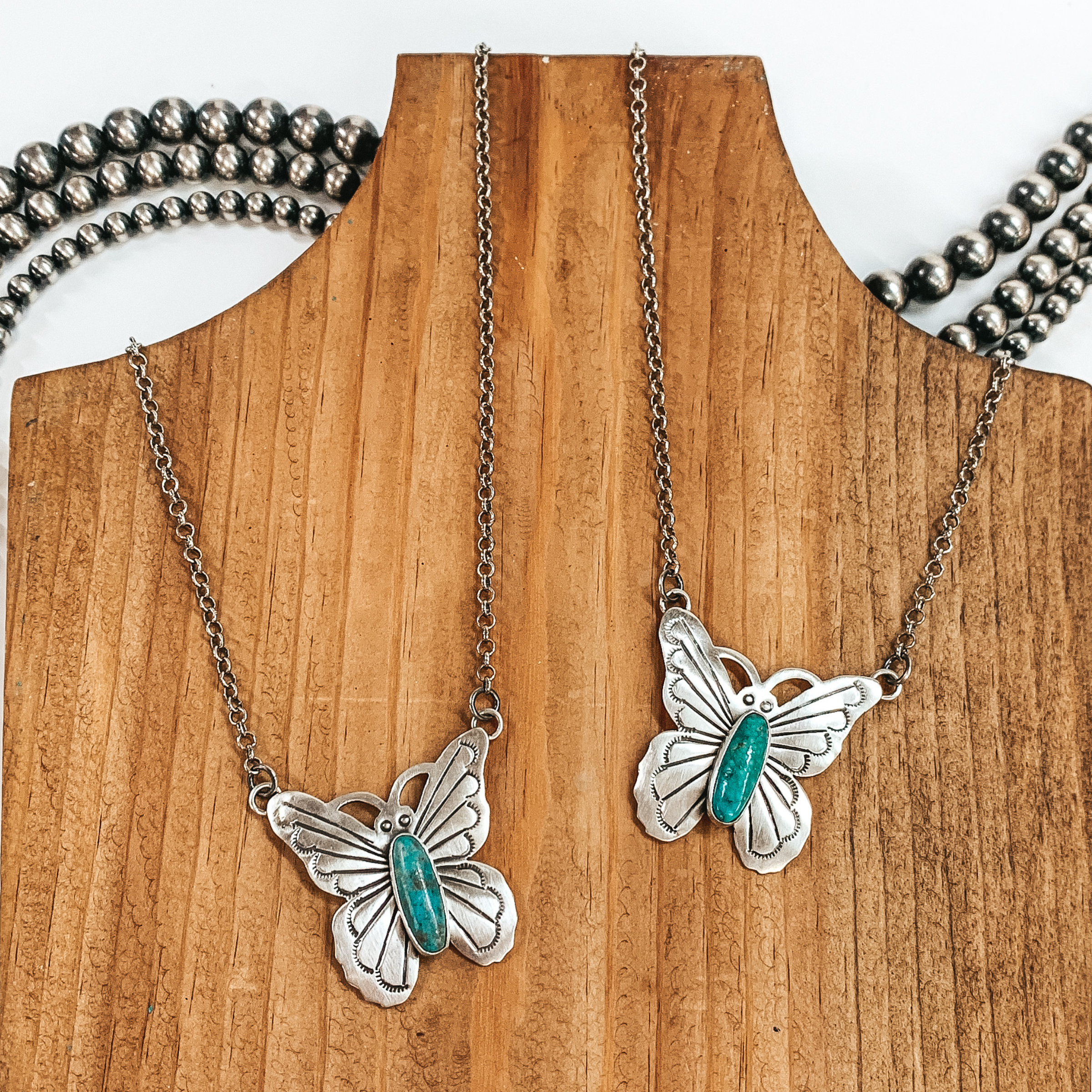 Two silver chain necklaces with a silver butterfly pendant. The pendant includes an engraved design and a center turquoise stone. These necklaces are pictured on a wooden necklace holder on a white background. 