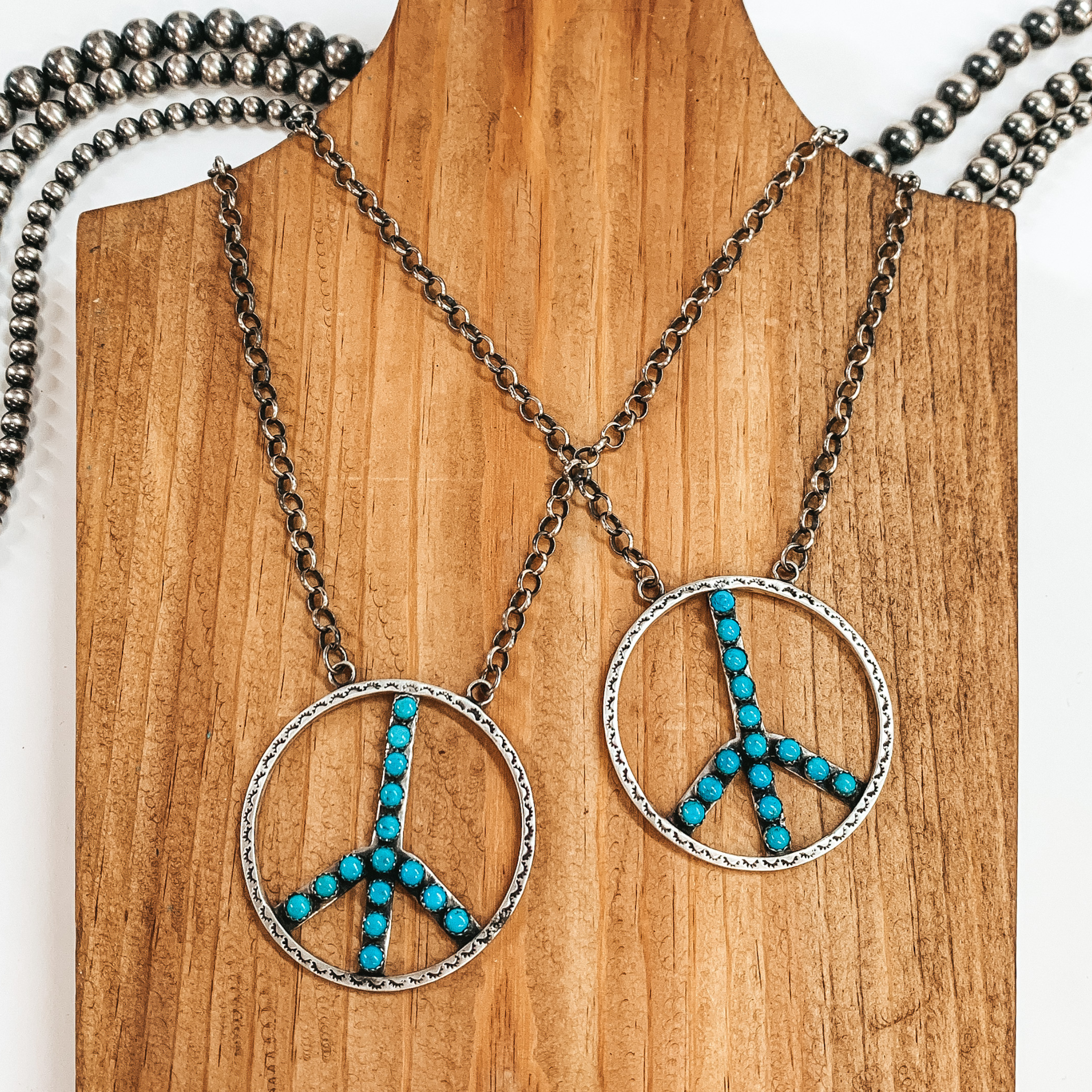 Two silver chain necklaces with a peace sign pendants. The pendants includes an engraved design and turquoise stone inlay. These necklaces are pictured on a wooden necklace holder on a white background.