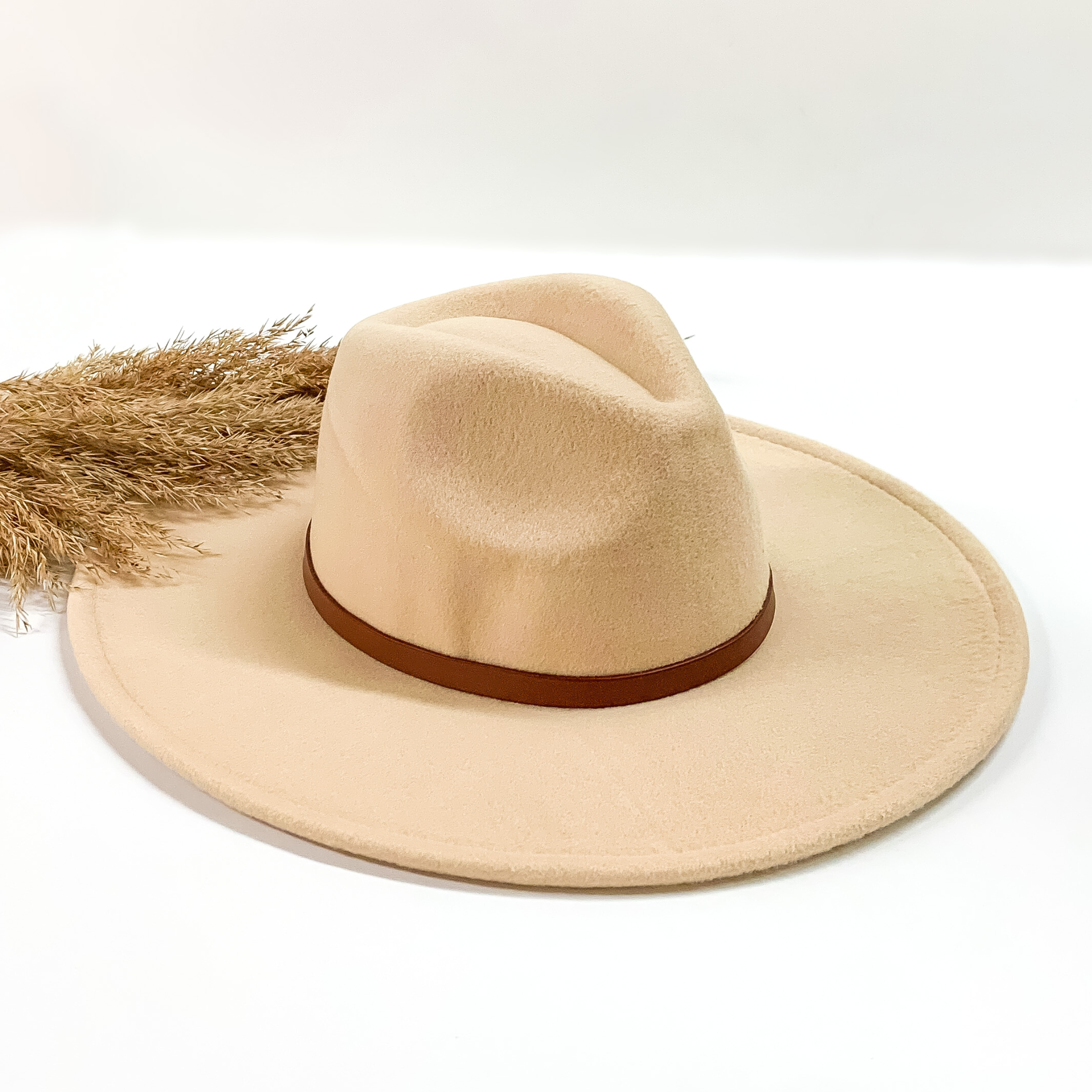 Ivory faux felt hat with a thin brown hat band. This hat is pictured on a white background with tan pompous in the background. 