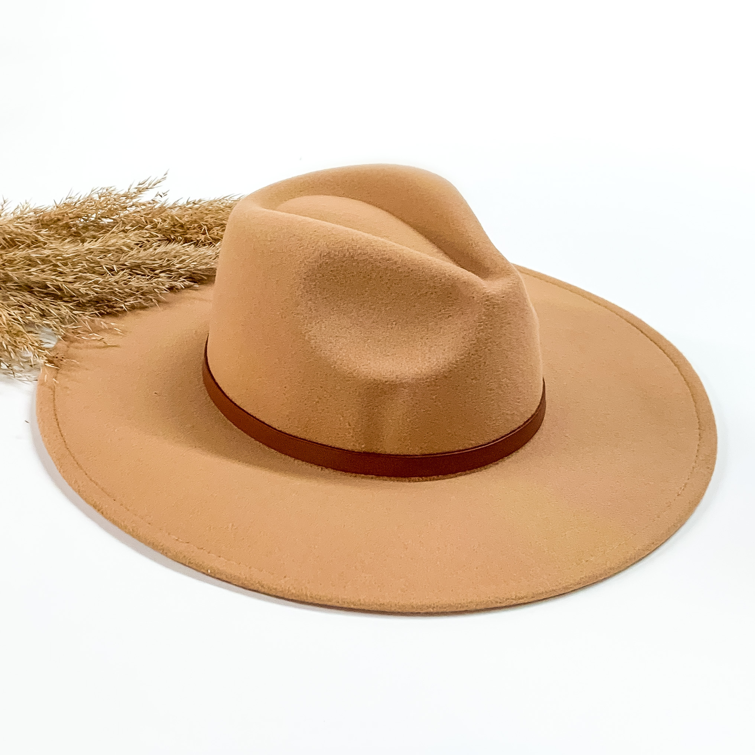 Tan faux felt hat with a thin brown hat band. This hat is pictured on a white background with tan pompous in the background. 