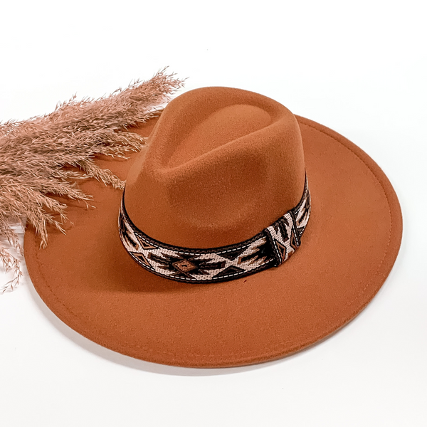 Rust faux felt hat with a aztec print embroidered hat band. This hat is pictured on a white background with tan pompous in the background. 
