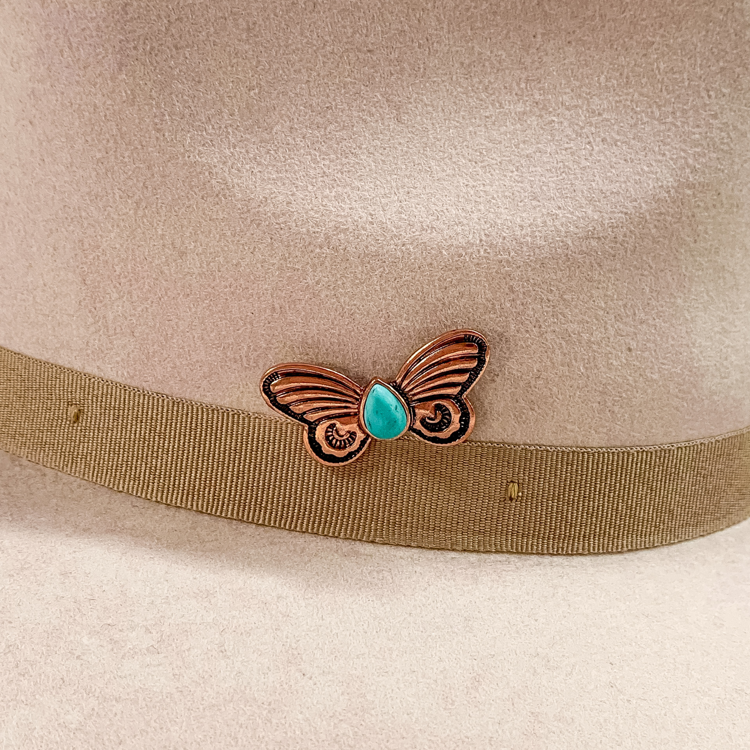 Copper butterfly hat pin with a center, teardrop, turquoise stone pictured on a beige colored hat pictured on a white background.