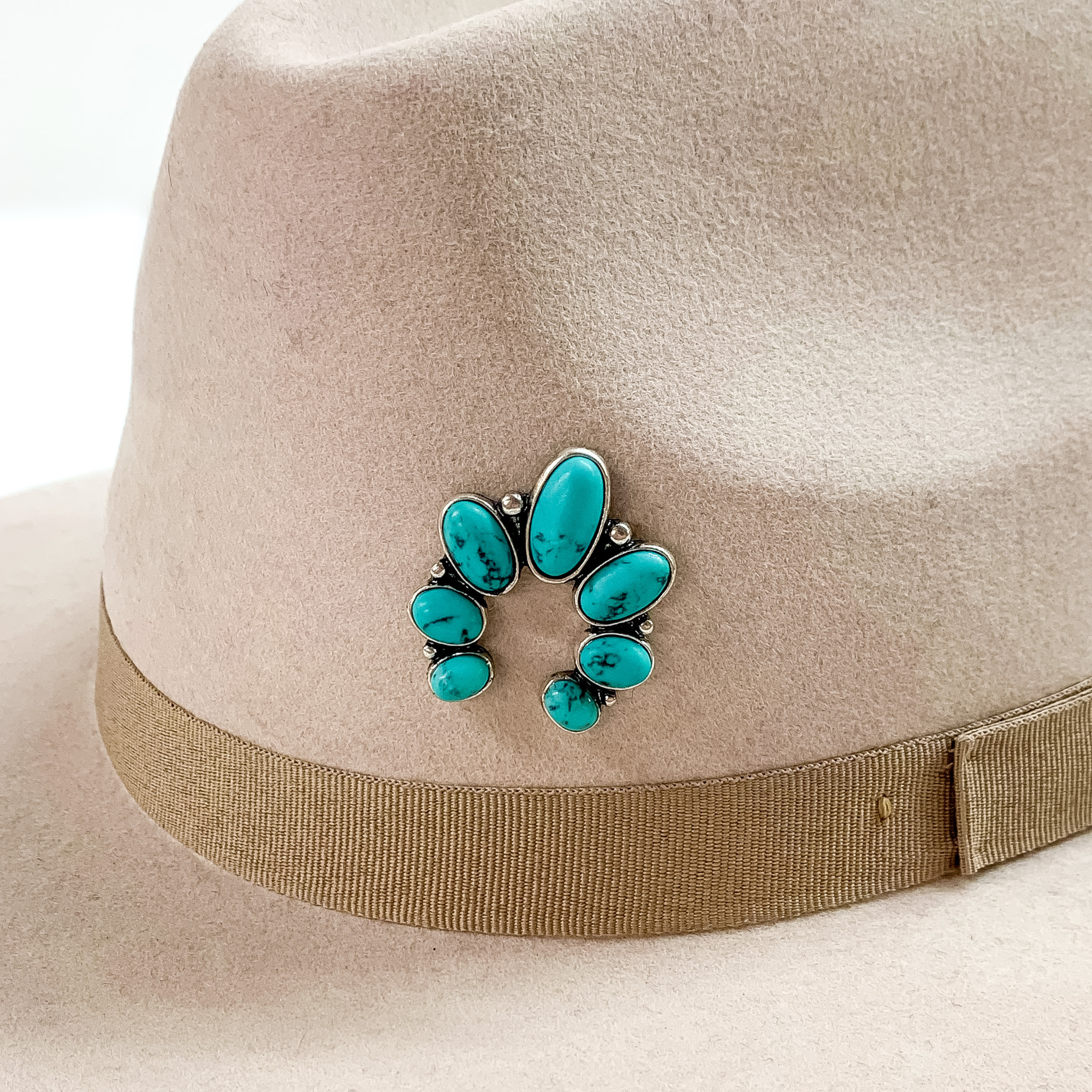 Silver naja hat pin with turquoise stones pictured on a beige colored hat pictured on a white background.