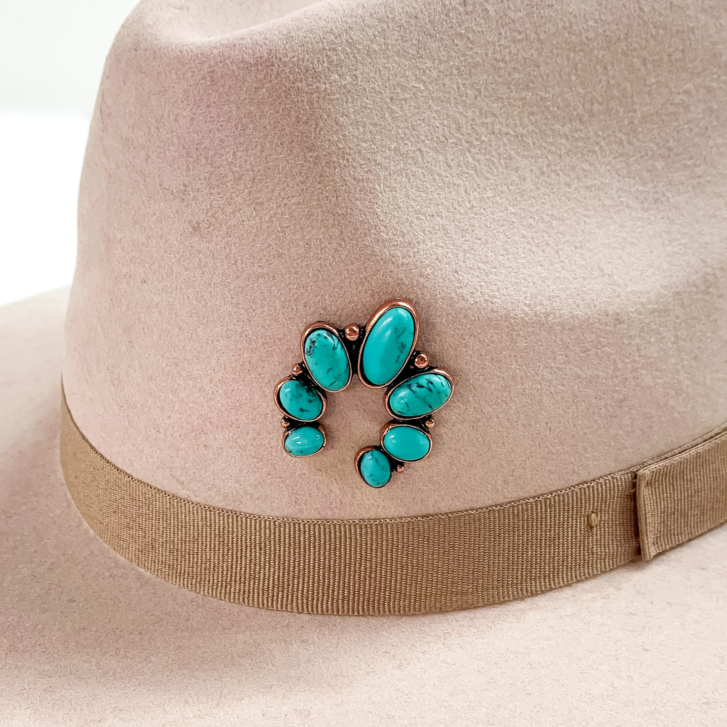 Copper naja hat pin with turquoise stones pictured on a beige colored hat pictured on a white background.