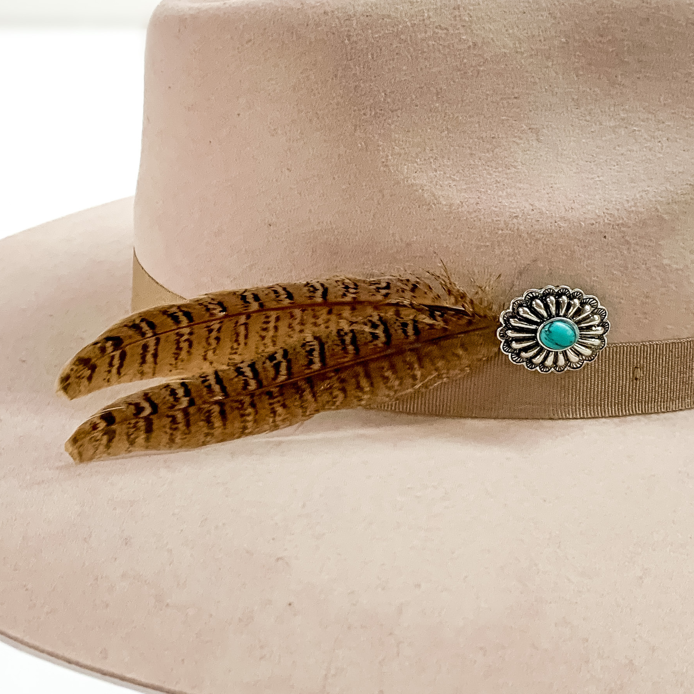 Silver, oval concho with center turquoise stone and two brown feathers. This hat pin is pictured on a beige colored hat on a white background.