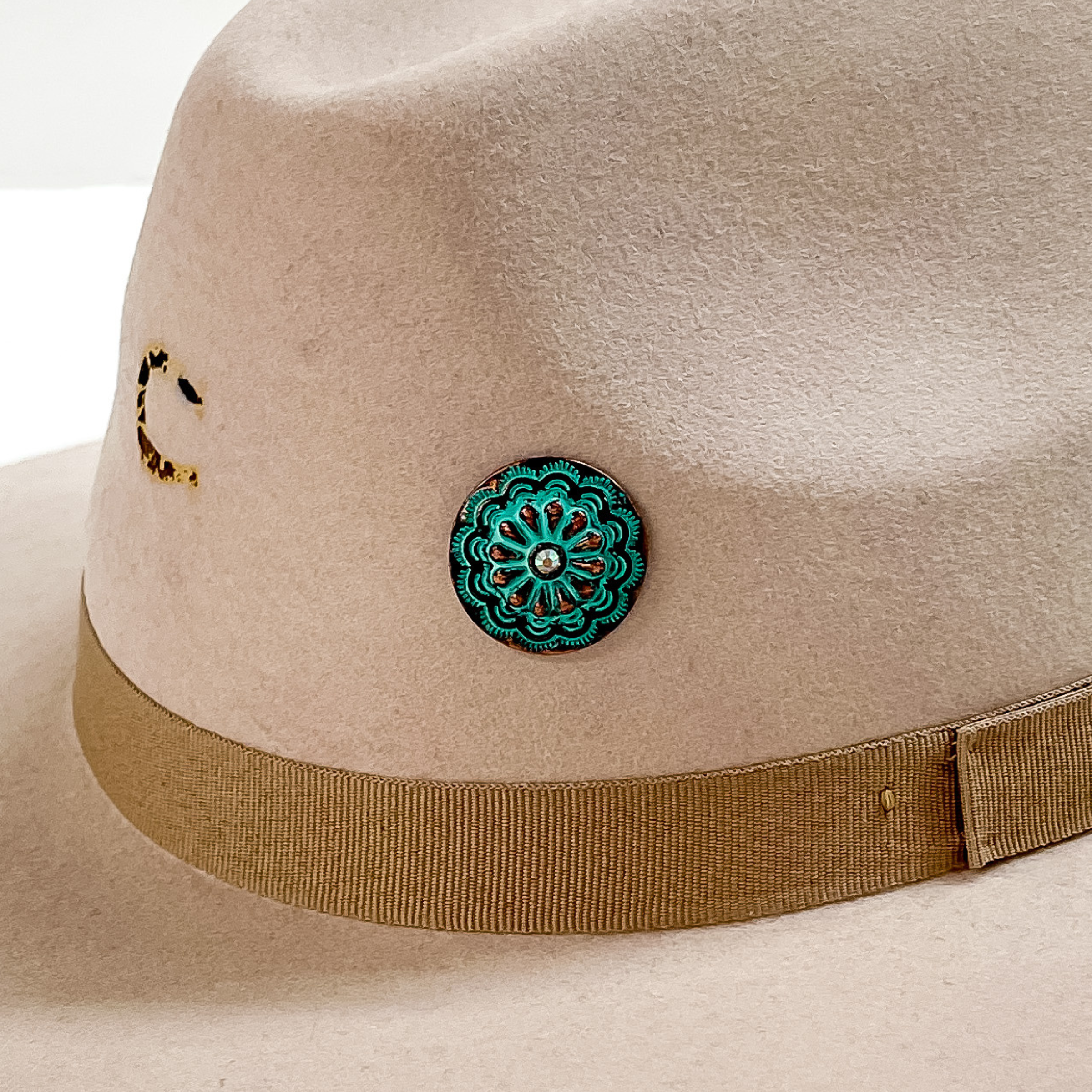 Patina, circle concho with detailed engraving and a small, center AB crystal hat pin. This hat pin is pictured on a beige hat and white background. 