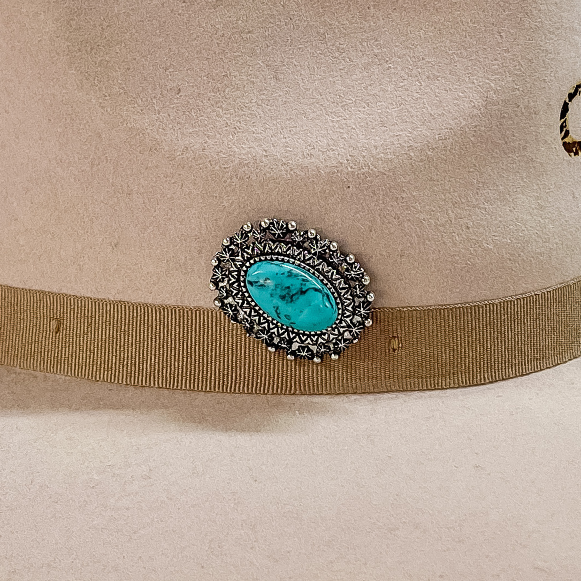 Silver, oval concho with detailed carving and a large center turquoise stone hat pin. This hat pin is pictured on a beige hat. 