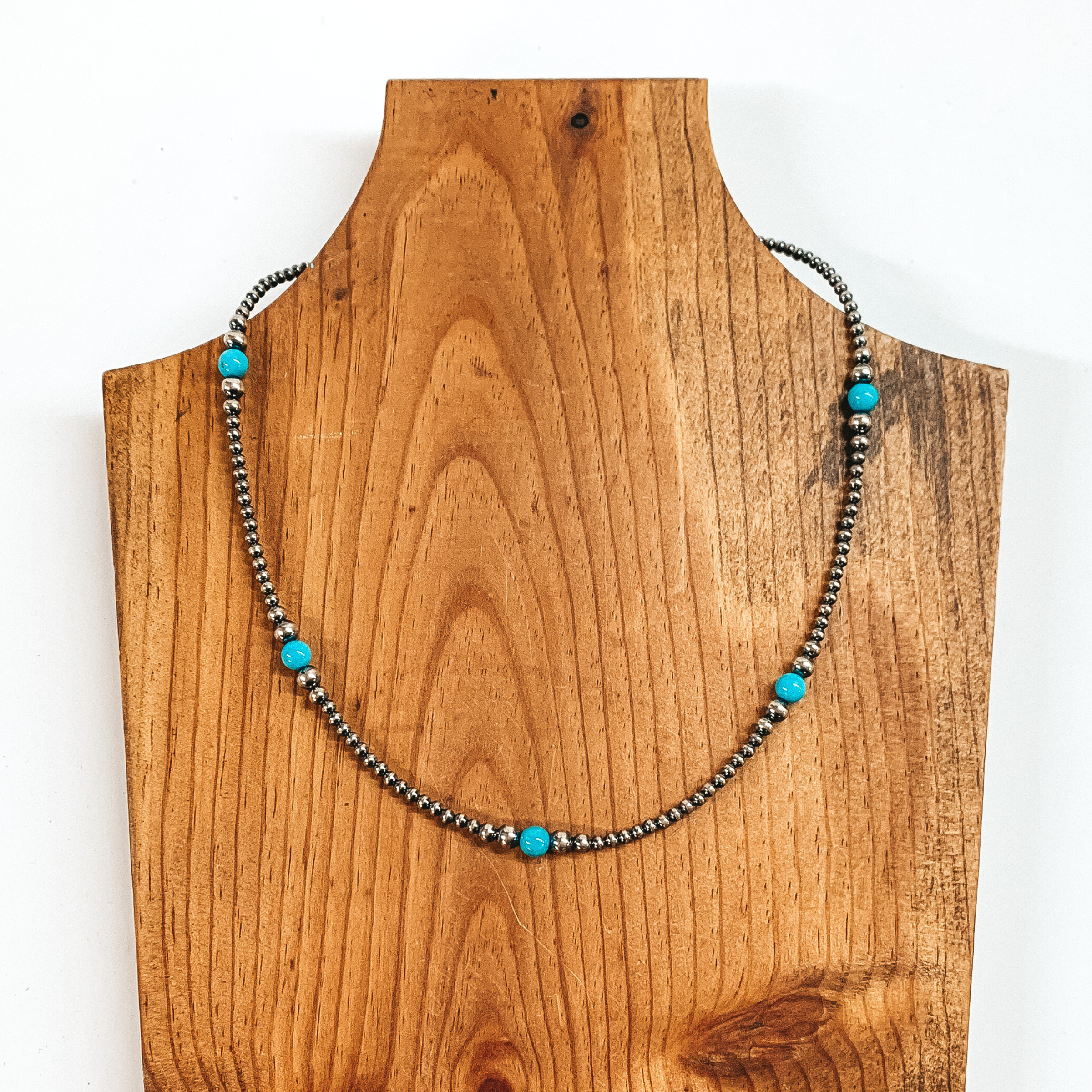 Silver beaded necklace with blue beaded spacers throughout. This necklace is pictured on a brown necklace holder on a white background. 