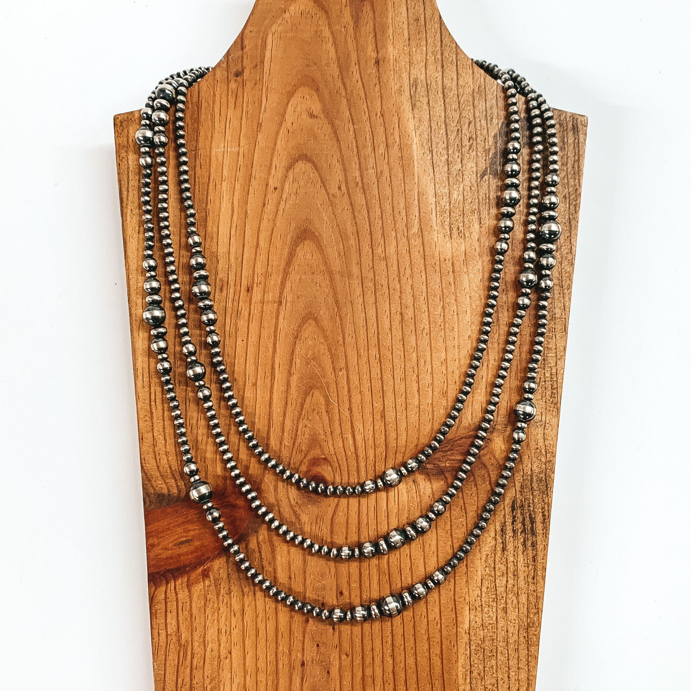 Three strand silver beaded necklace with different design and size beads through out. This necklace is pictured on a brown necklace holder on a white background. 