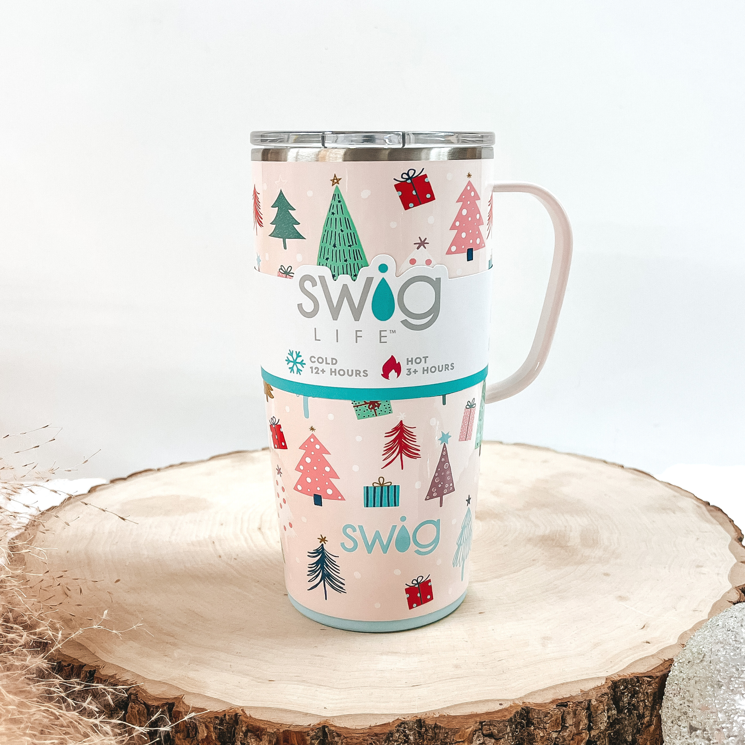 Blush pink mug with multicolored tree and present print with a blush pink handle and clear lid. This can cooler is pictured on a piece of wood on a white background.