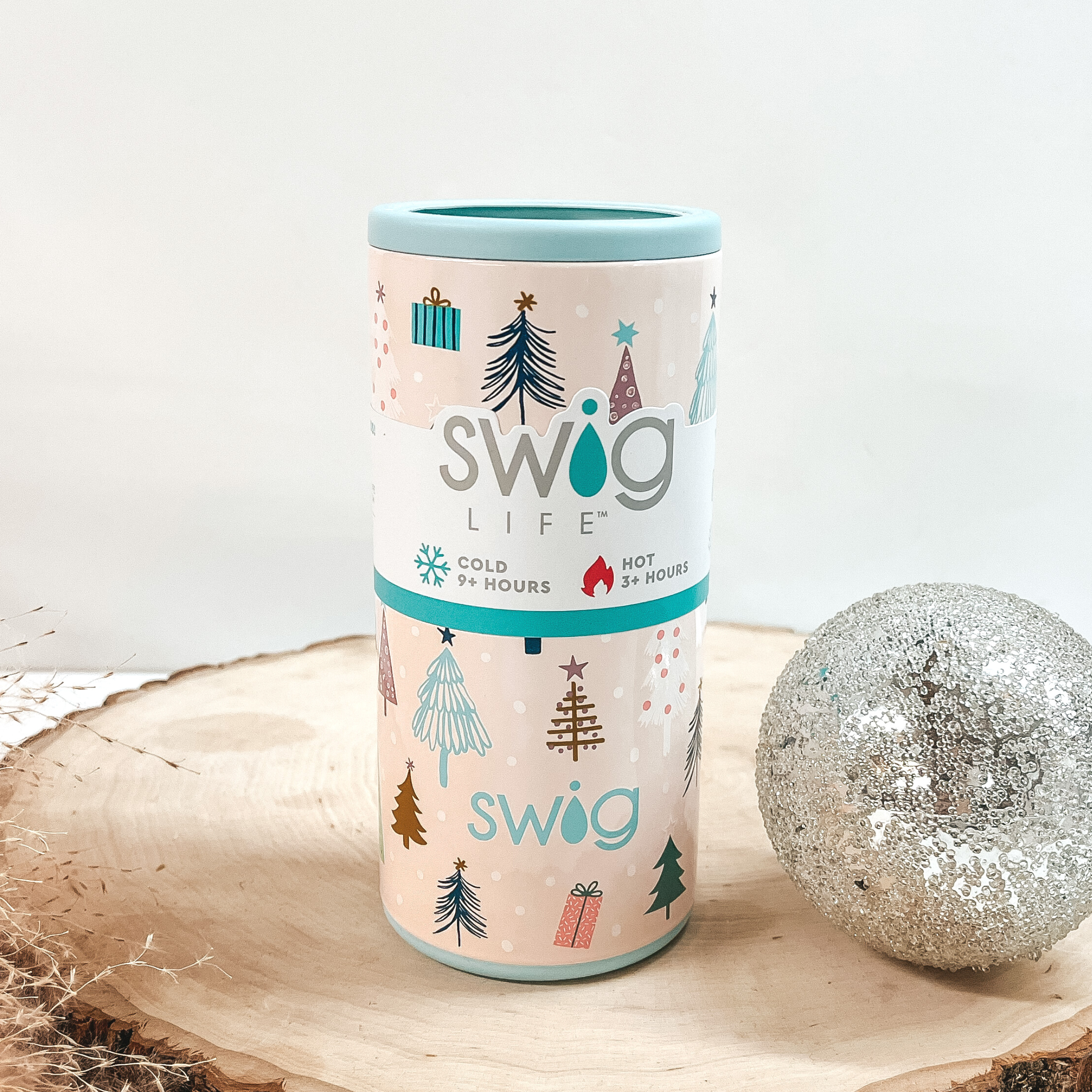 A blush pink slim can cooler with multicolored tree and present print. This can cooler is pictured on a piece of wood on a white background with a silver ball next to it. 