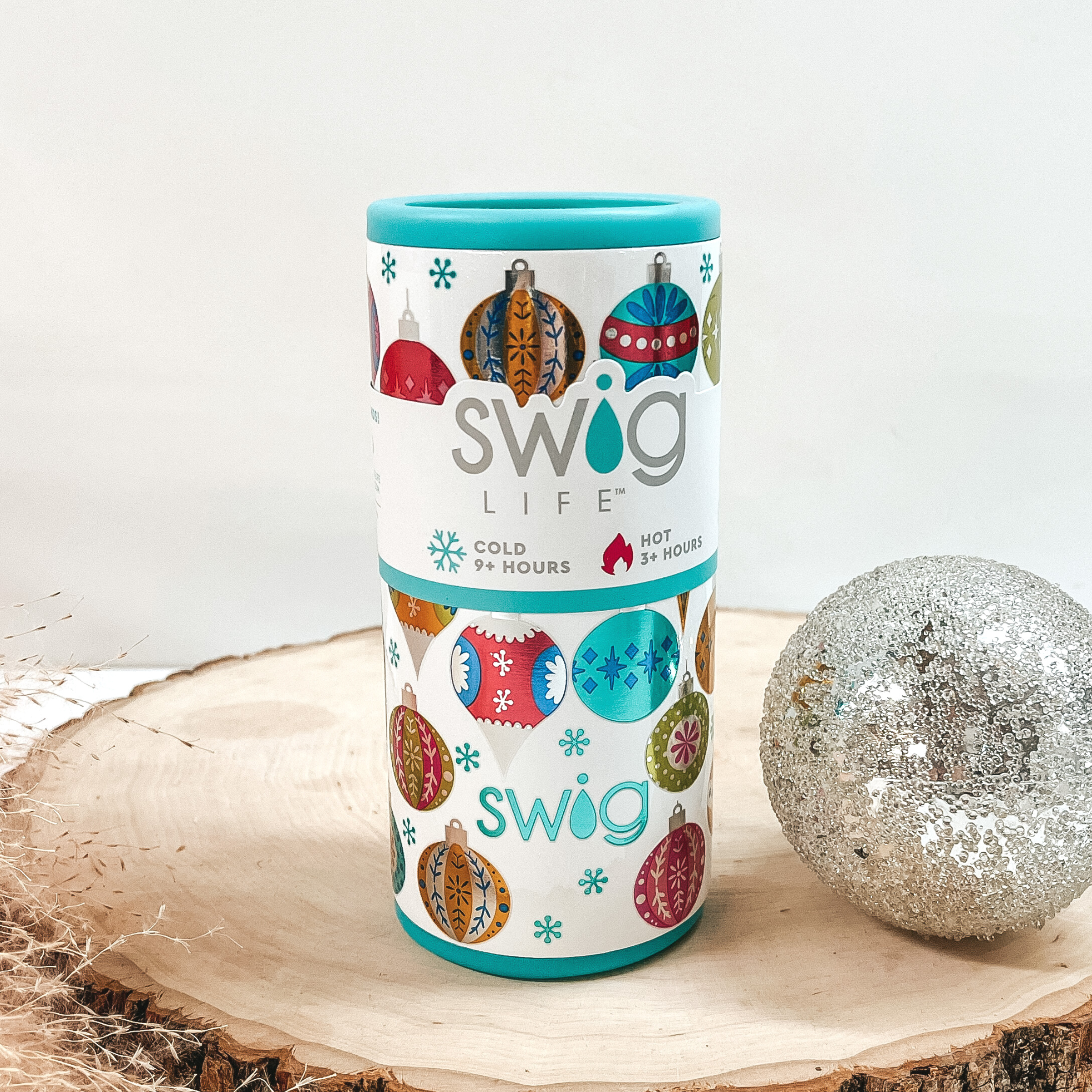 A white slim can cooler with shiny, multicolored ornament print with blue snowflakes throughout. This can cooler is pictured on a piece of wood on a white background with a silver ball next to it. 