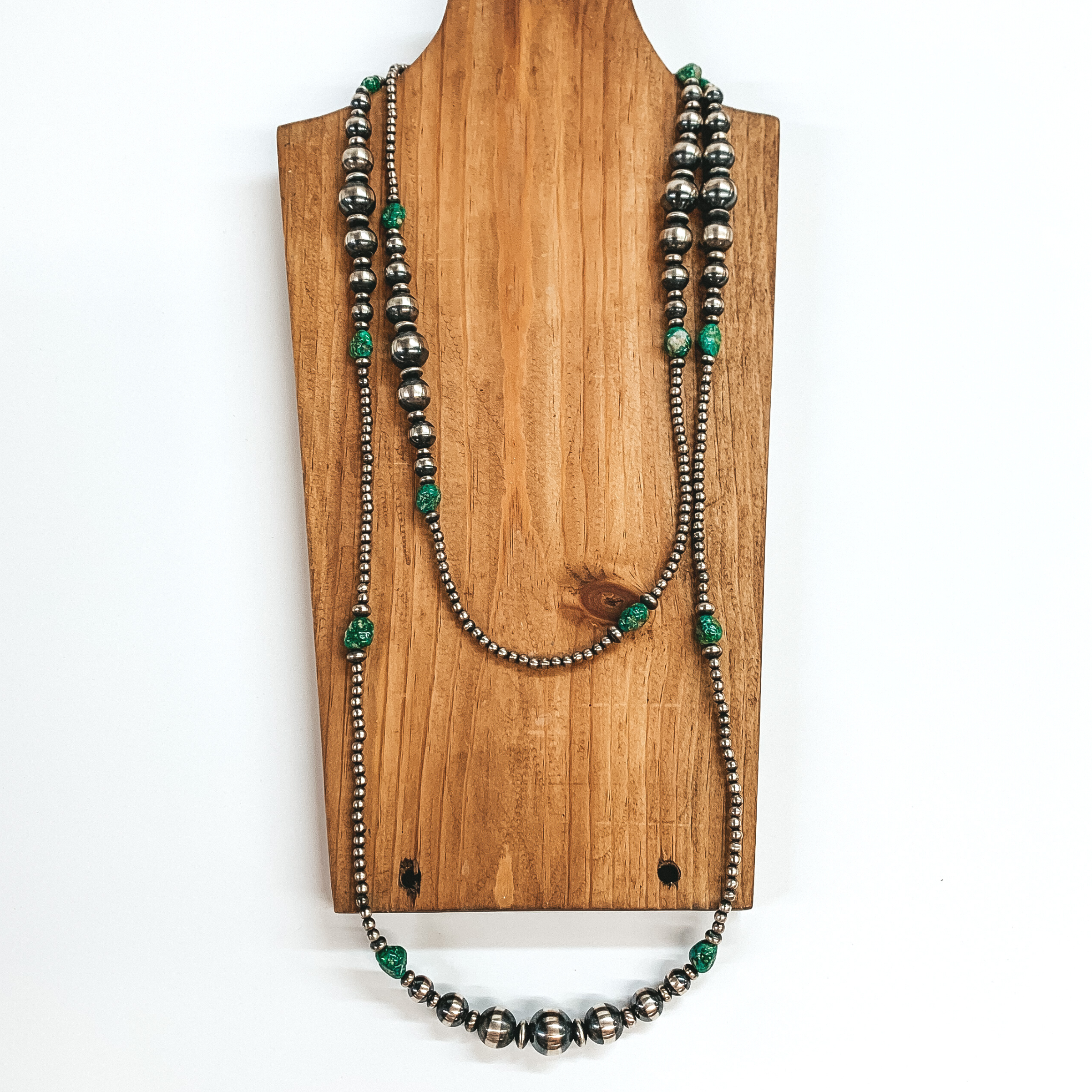 Silver graduated beaded necklace with green stone spacers. This necklace is pictured laying on a wood necklace holder on a white background. 