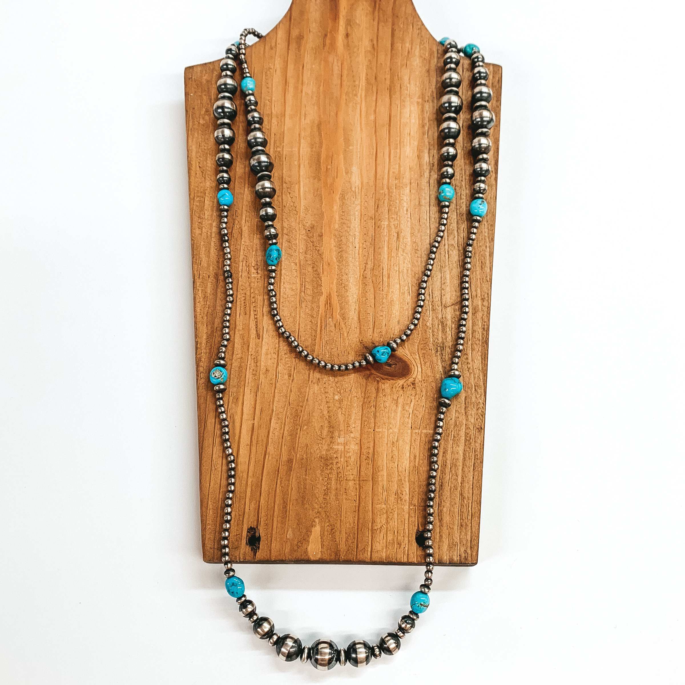 Silver graduated beaded necklace with turquoise stone spacers. This necklace is pictured laying on a wood necklace holder on a white background.