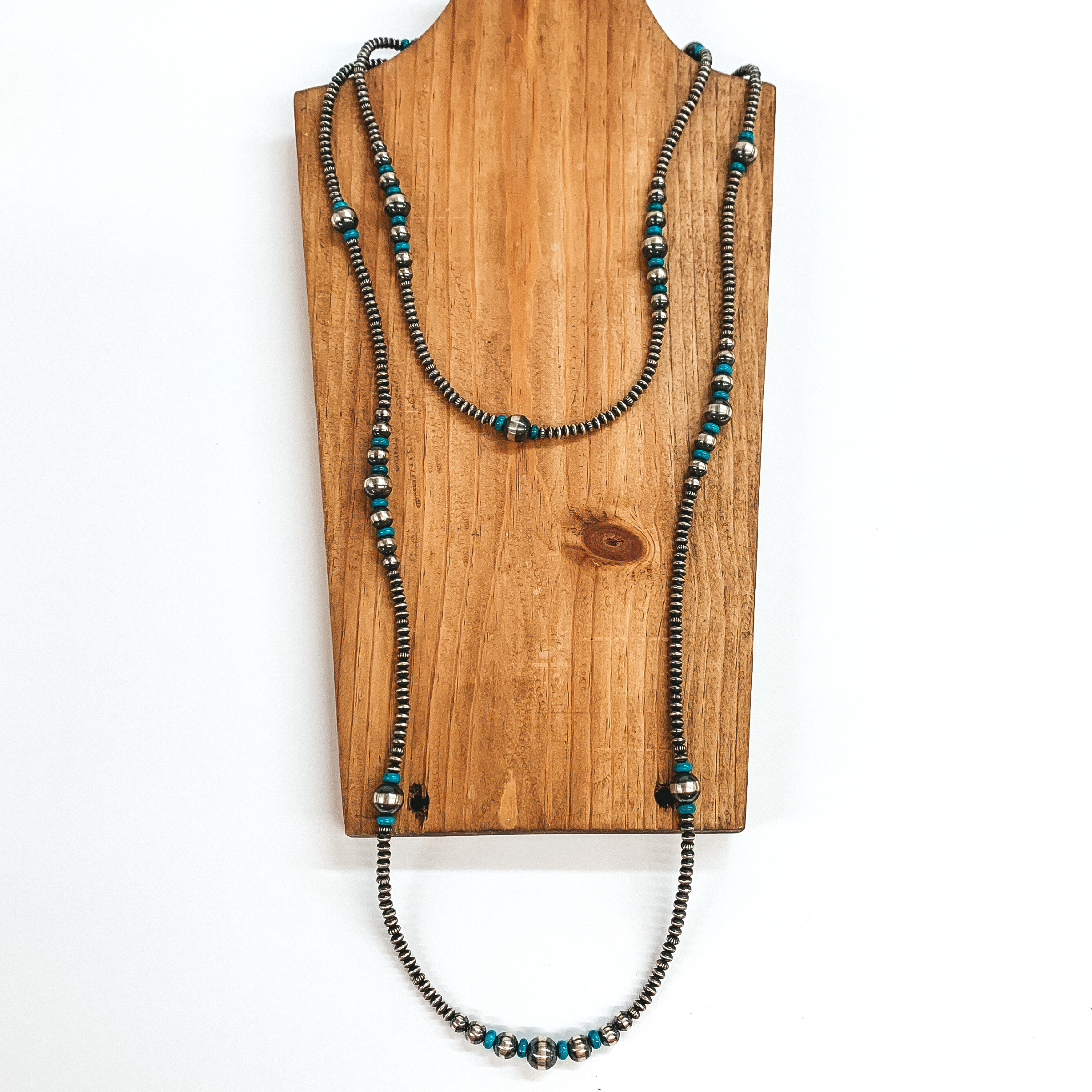 Silver saucer beaded necklace with turquoise stone spacers and graduated silver beads. This necklace is pictured laying on a wood necklace holder on a white background.