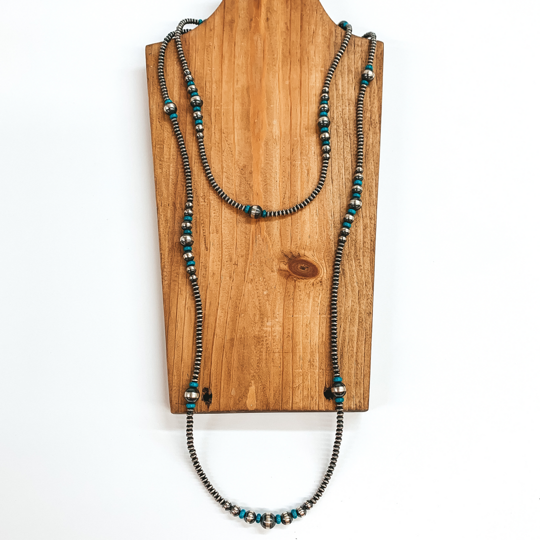 Silver saucer beaded necklace with turquoise stone spacers and graduated silver beads. This necklace is pictured laying on a wood necklace holder on a white background.