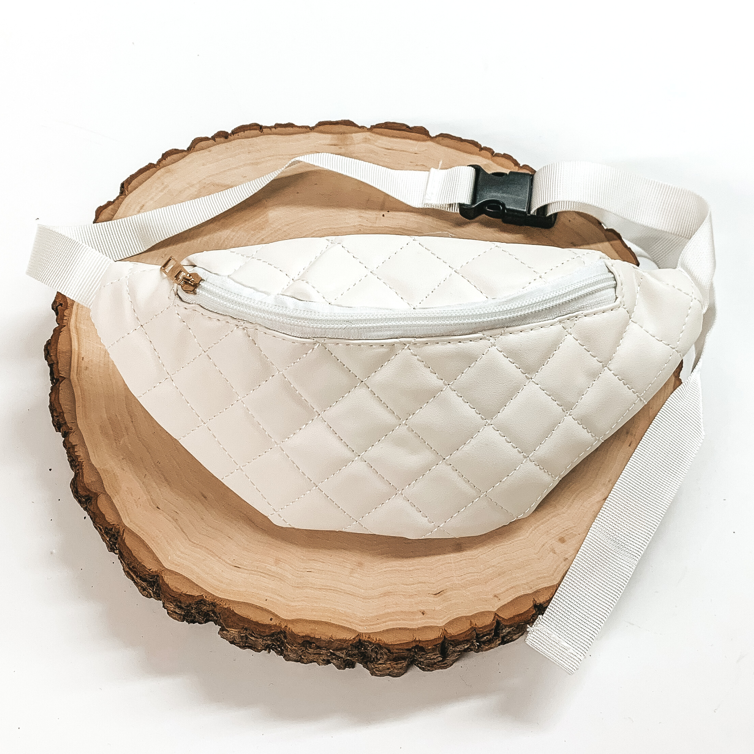 White, quilted fanny pack with a zipper across the front. This fanny pack also includes white straps and black clips. This fanny pack is pictured on a piece of wood on a white background. 