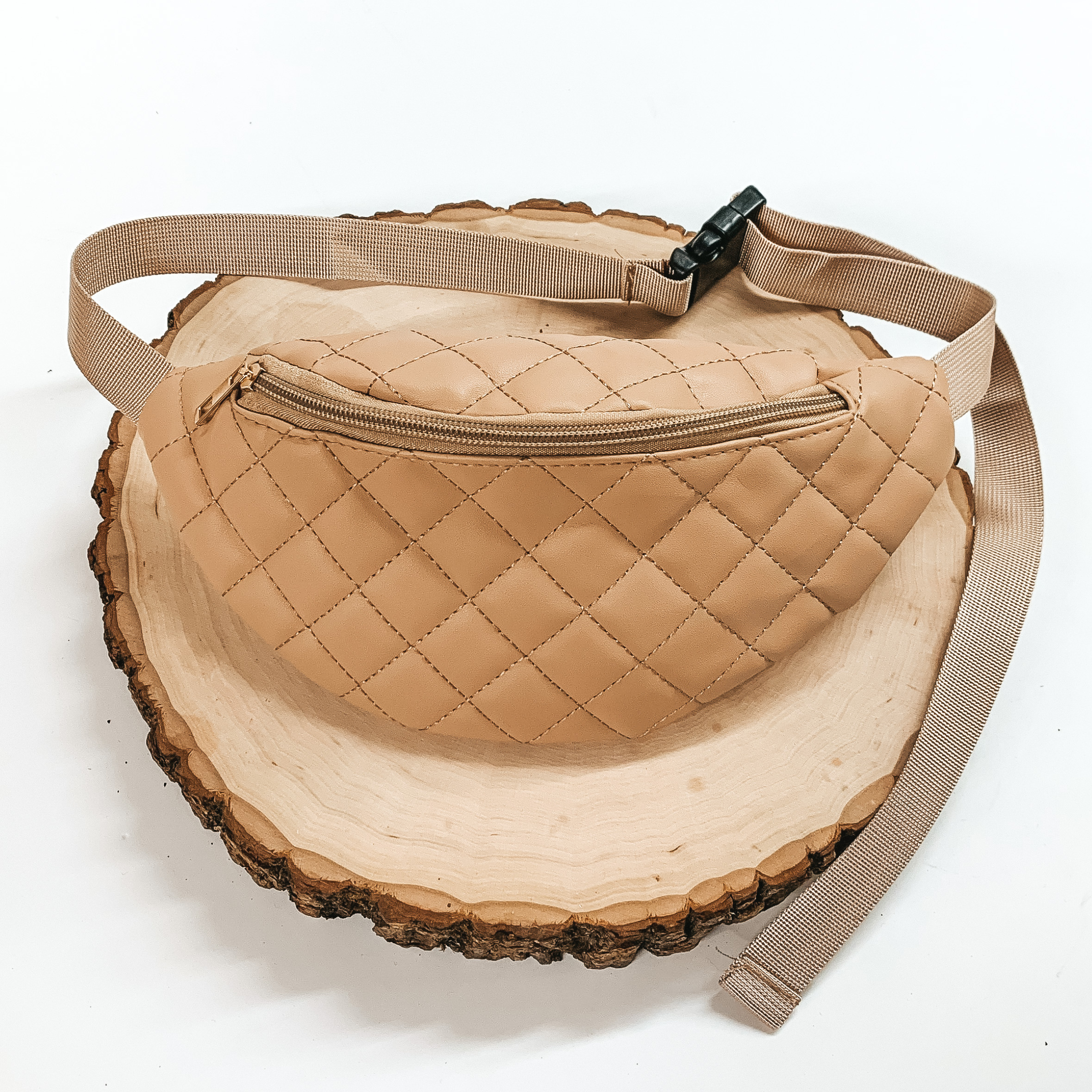 Beige, quilted fanny pack with a zipper across the front. This fanny pack also includes beige straps and black clips. This fanny pack is pictured on a piece of wood on a white background. 