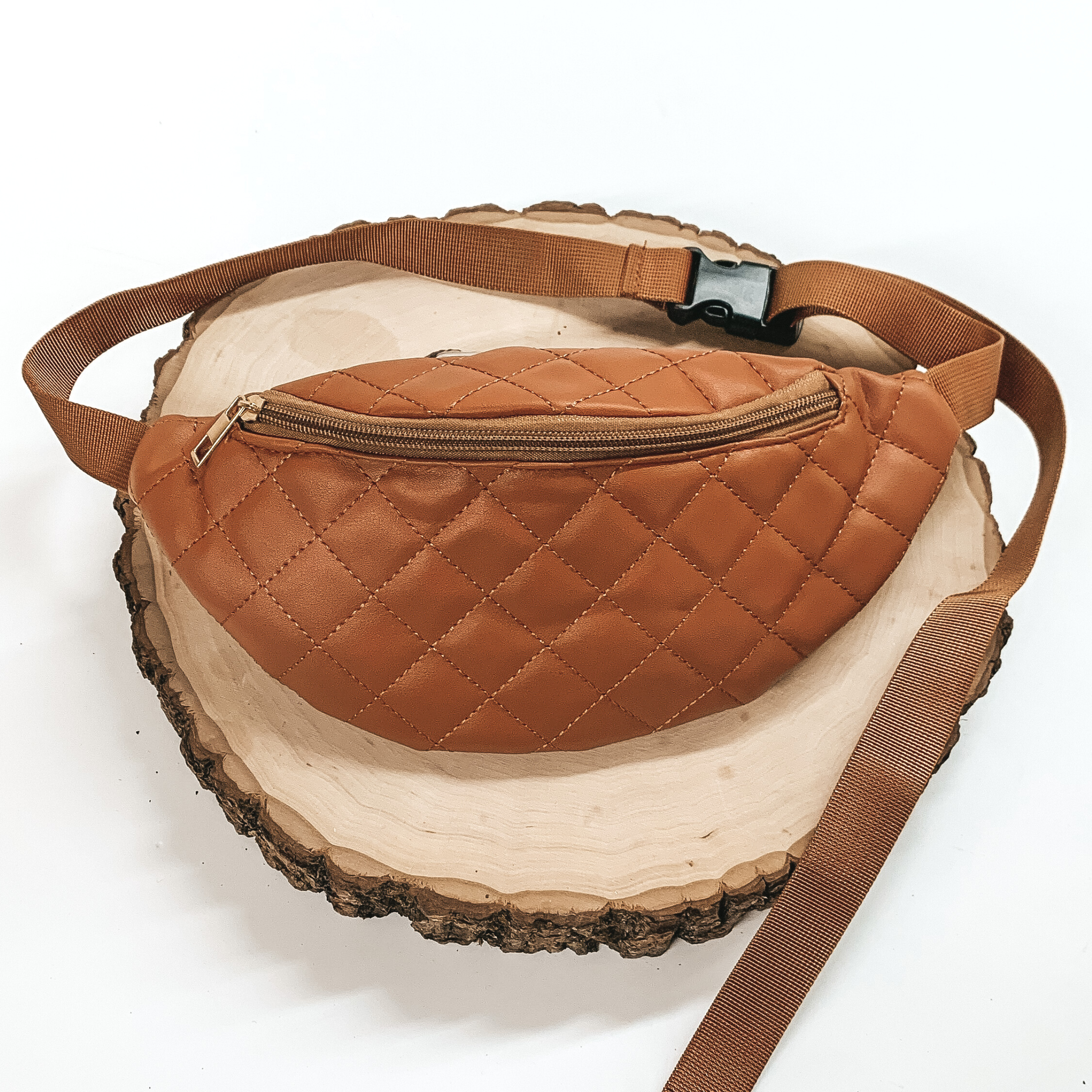 Cognac, quilted fanny pack with a zipper across the front. This fanny pack also includes brown straps and black clips. This fanny pack is pictured on a piece of wood on a white background. 