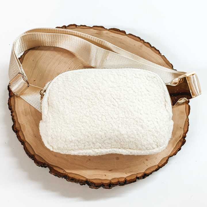 Ivory, sherpa fanny pack with a top zipper across the front. This fanny pack also includes ivory straps and gold clips. This fanny pack is pictured on a piece of wood on a white background. 