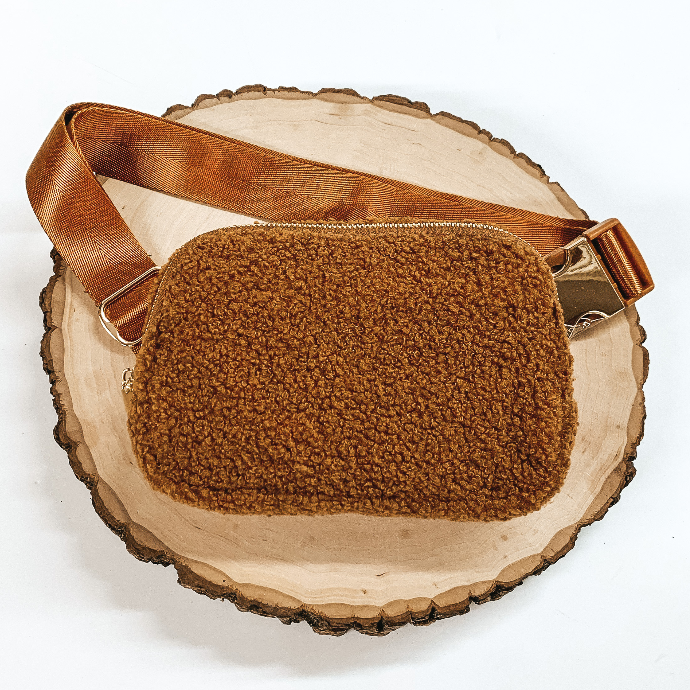 Brown, sherpa fanny pack with a top zipper across the front. This fanny pack also includes brown straps and gold clips. This fanny pack is pictured on a piece of wood on a white background. 