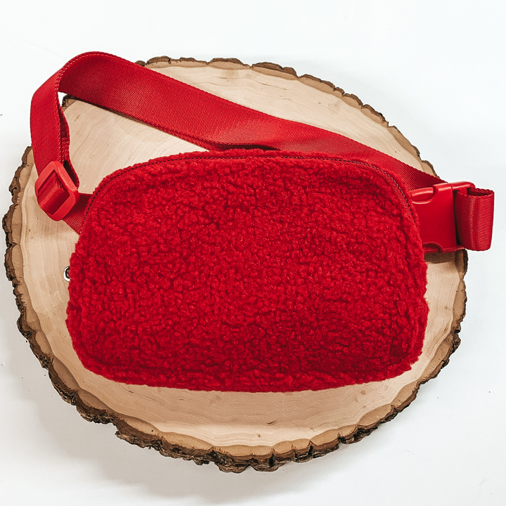 Red, sherpa fanny pack with a top zipper across the front. This fanny pack also includes red straps and red clips. This fanny pack is pictured on a piece of wood on a white background. 