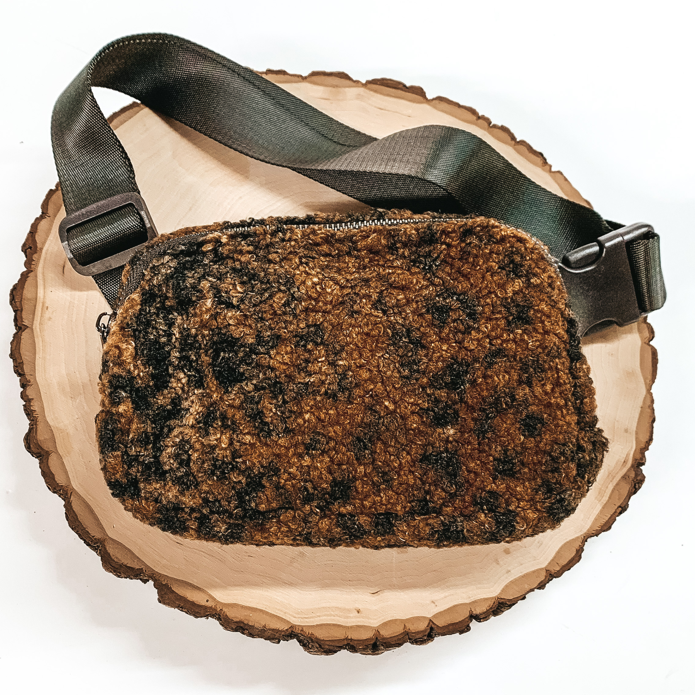 Leopard print, sherpa fanny pack with a top zipper across the front. This fanny pack also includes dark brown straps and dark brown clips. This fanny pack is pictured on a piece of wood on a white background. 