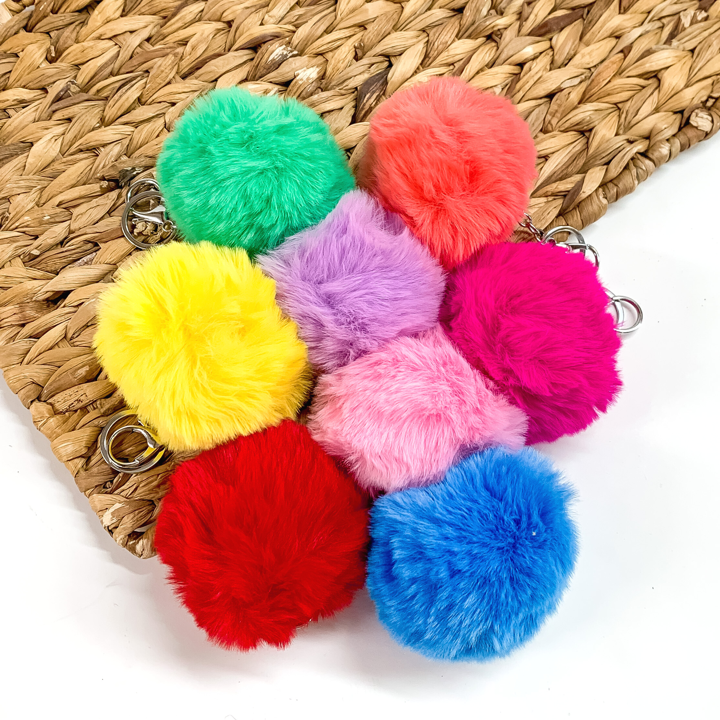 There are eight different colored puff ball keychains with a silver chain. There is green, coral, yellow, light purple, hot pink, light pink, red, and blue. These puff ball keychains are taken partly on a brown woven slab and on a white background.