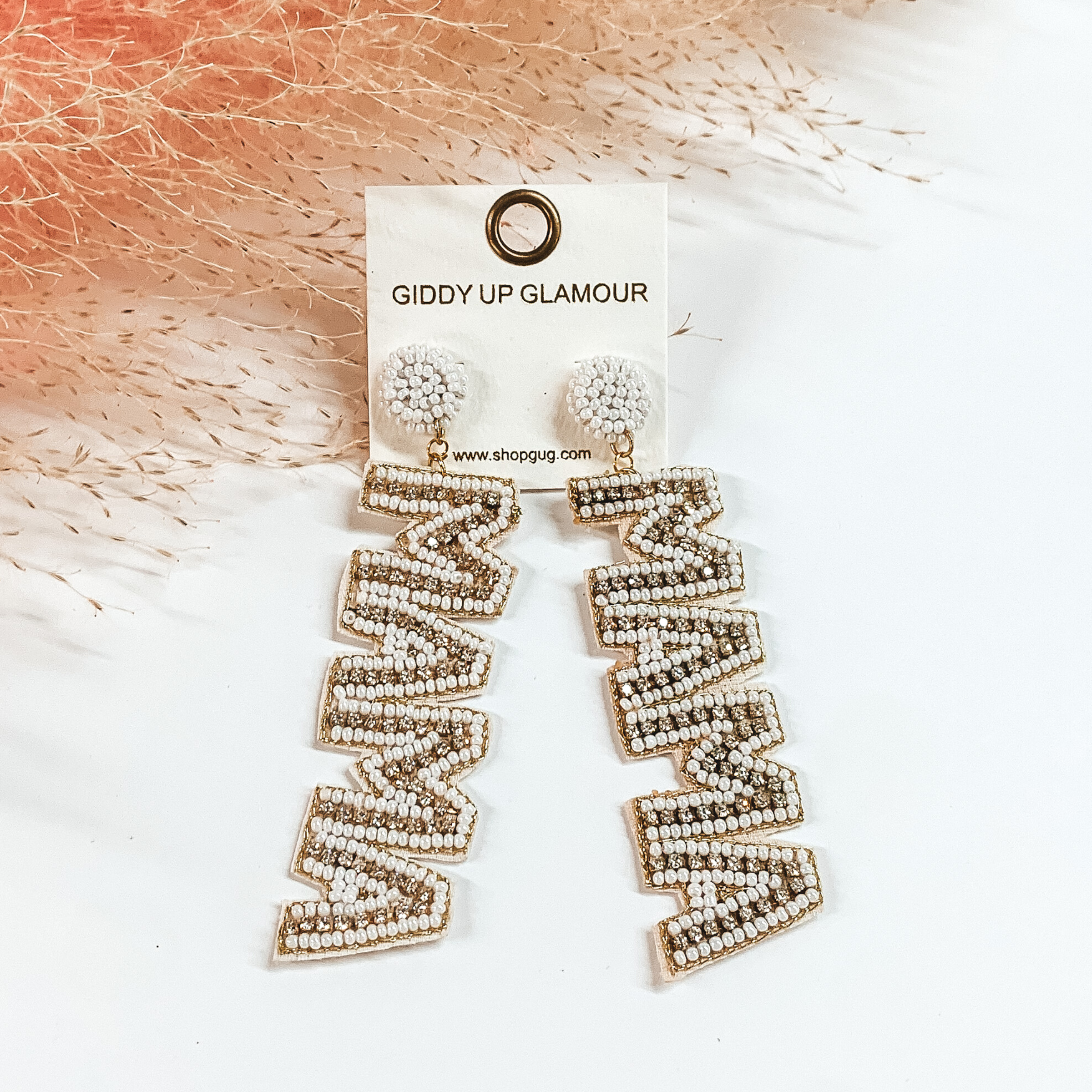 These earrings include white, circle, beaded post back earrings with a long dangle. This dangle spells out "MAMA" in white beads with an inlay of clear crystals. These earrings are pictured on a white background with pink pompous grass at the top. 
