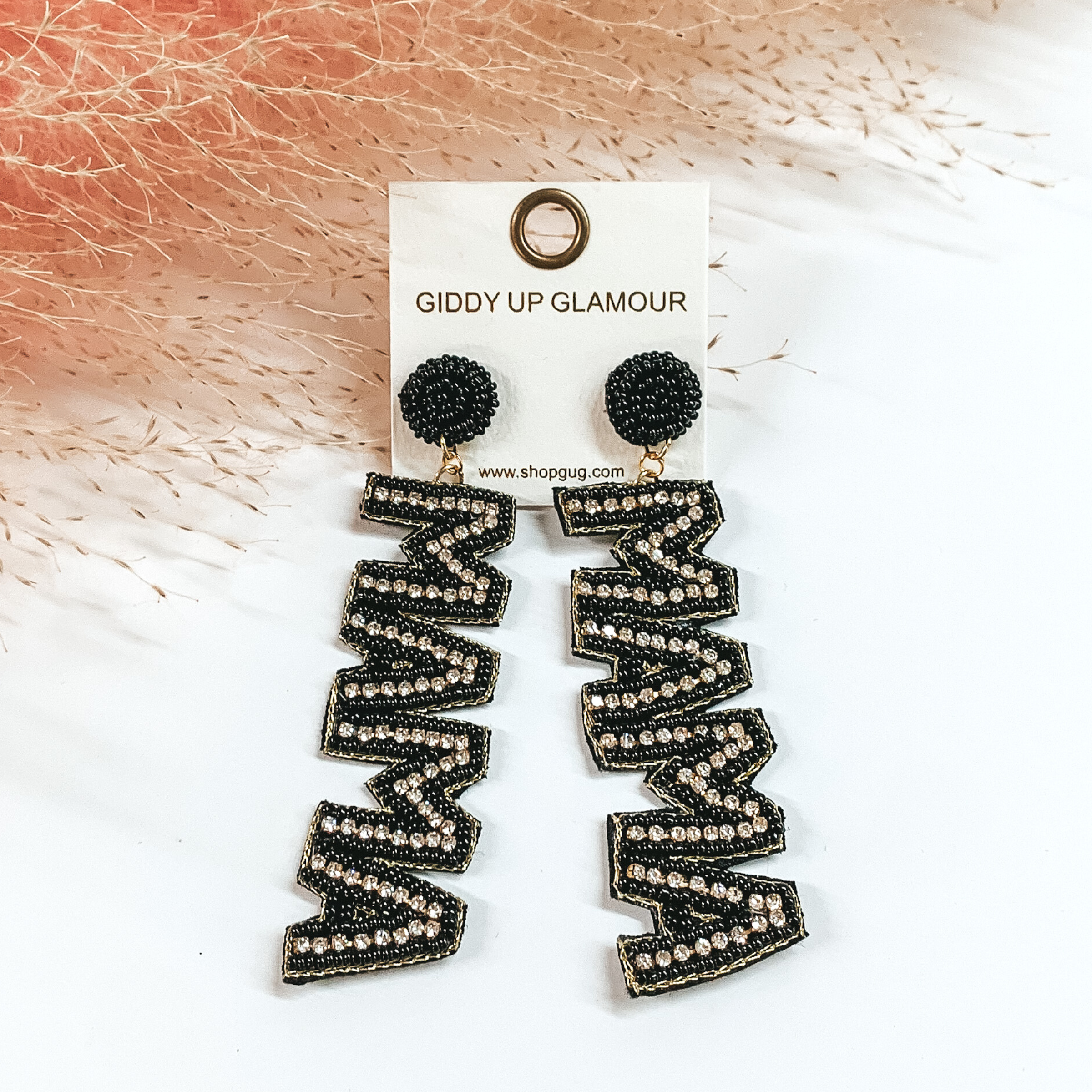 These earrings include black, circle, beaded post back earrings with a long dangle. This dangle spells out "MAMA" in black beads with an inlay of clear crystals. These earrings are pictured on a white background with pink pompous grass at the top. 