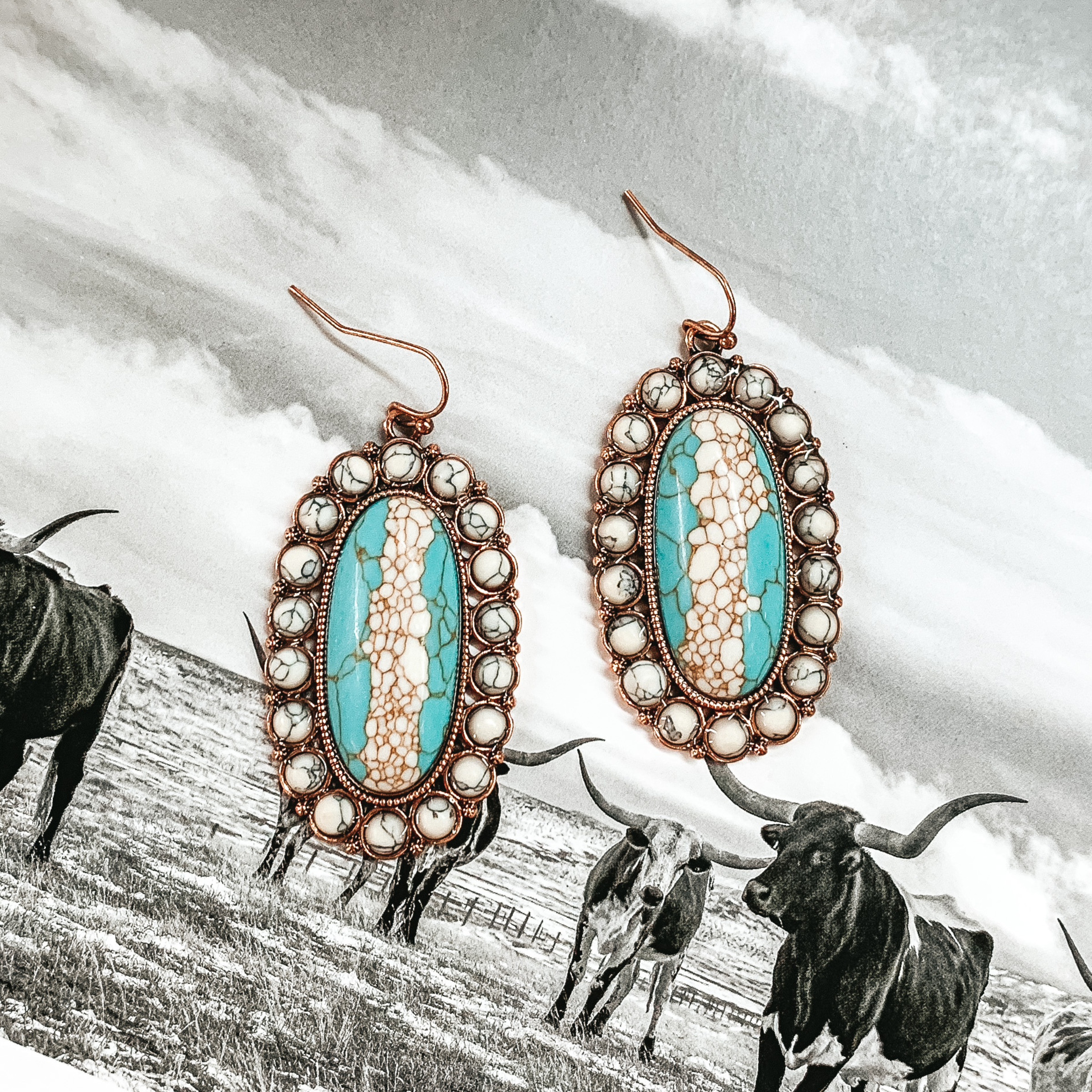 These oval, stone cluster earrings with a large center stone and smaller stones outlining it. The colors include turquoise and white. These earrings are pictured on a black and white picture of longhorns. 