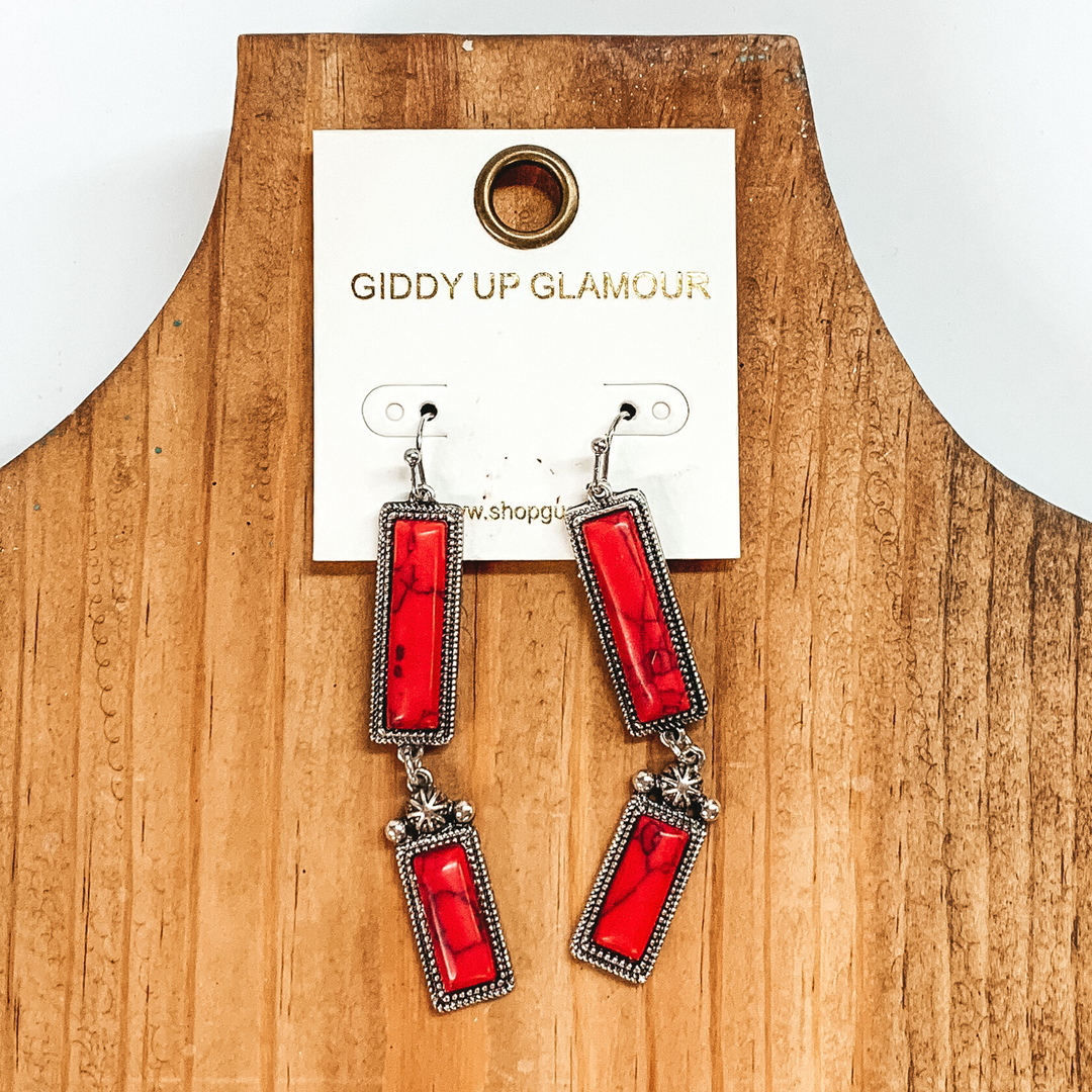 Silver drop earrings with two silver, rectangle drop. These earrings also include faux red stones. These earrings are pictured on a white and black picture. 