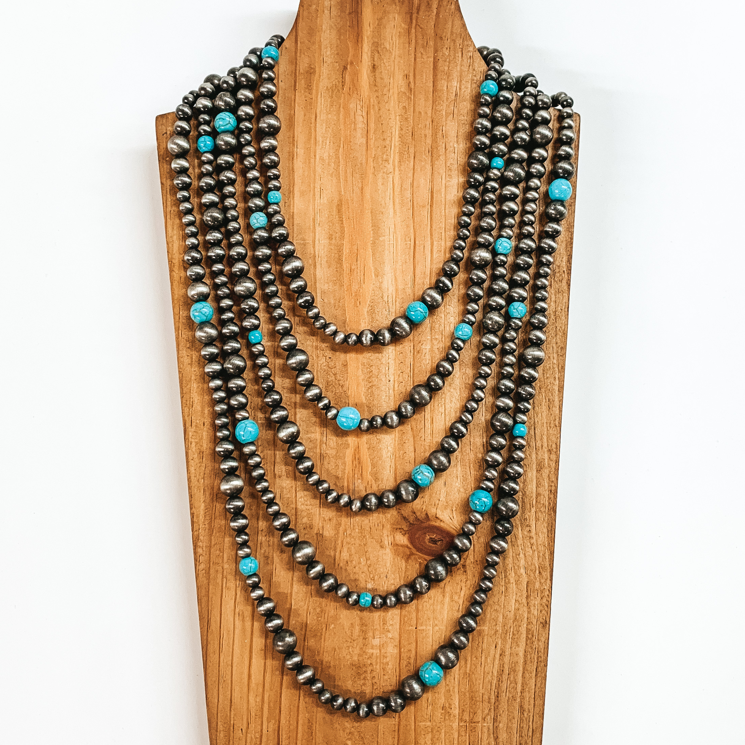 Five strand necklace that is mostly silver beads with a some turquoise bead spacers. This necklace is pictured on a wooden necklace holder on a white background. 