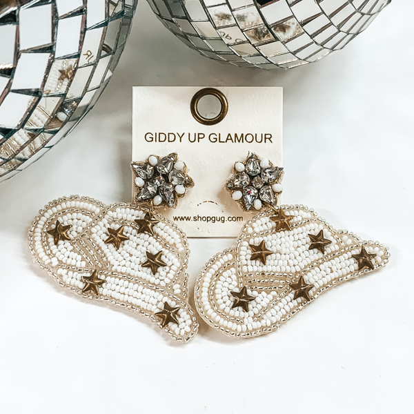 White beaded cowboyd hat earrings that include gold star charms. These earrings are pictured on a white background with disco balls at the top. 