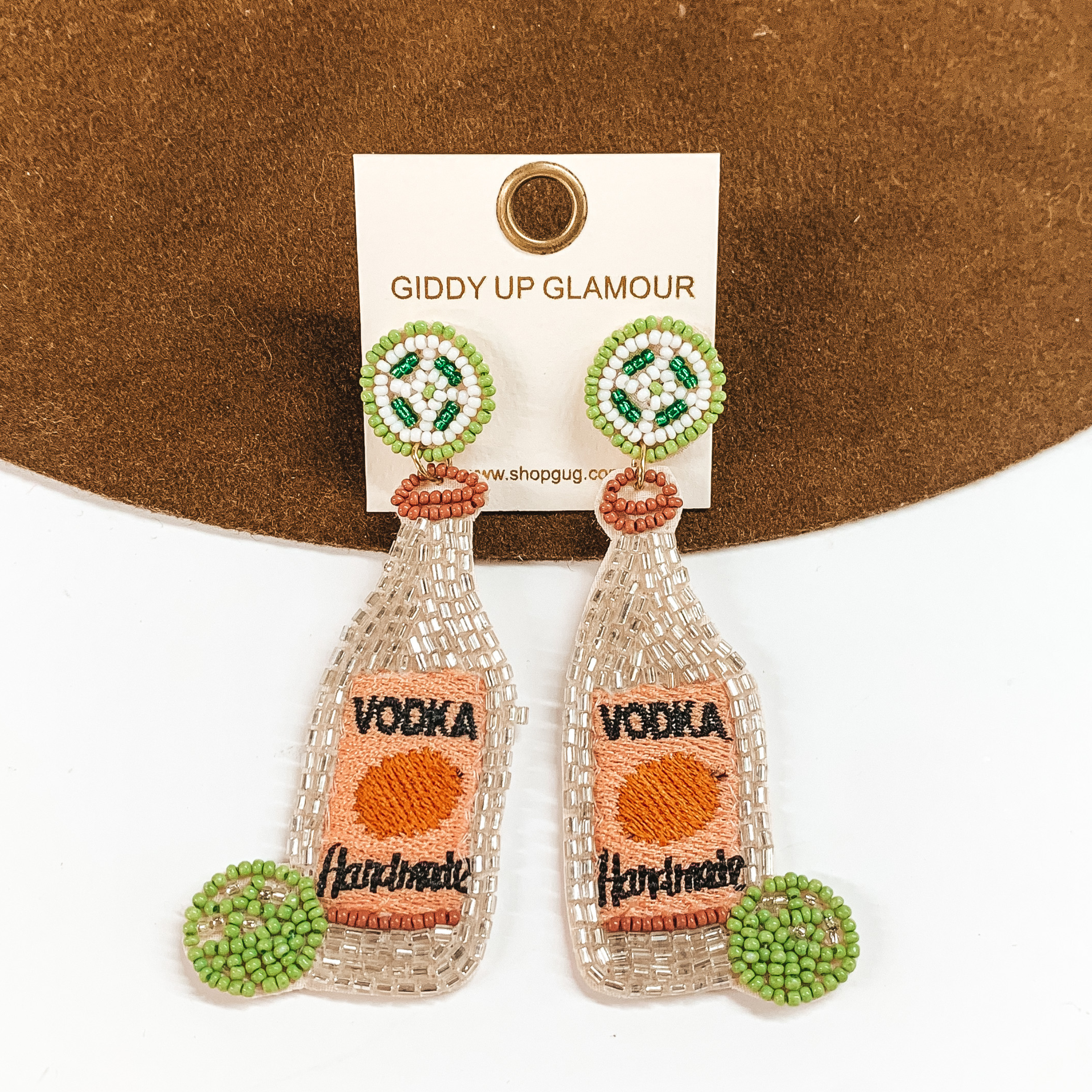 White beaded bottle earrings with lime beaded post studs. These earrings aso include a black stitching that spells out "VODKA" and "Handmade". These earrings are pictured on a white and brown background. 