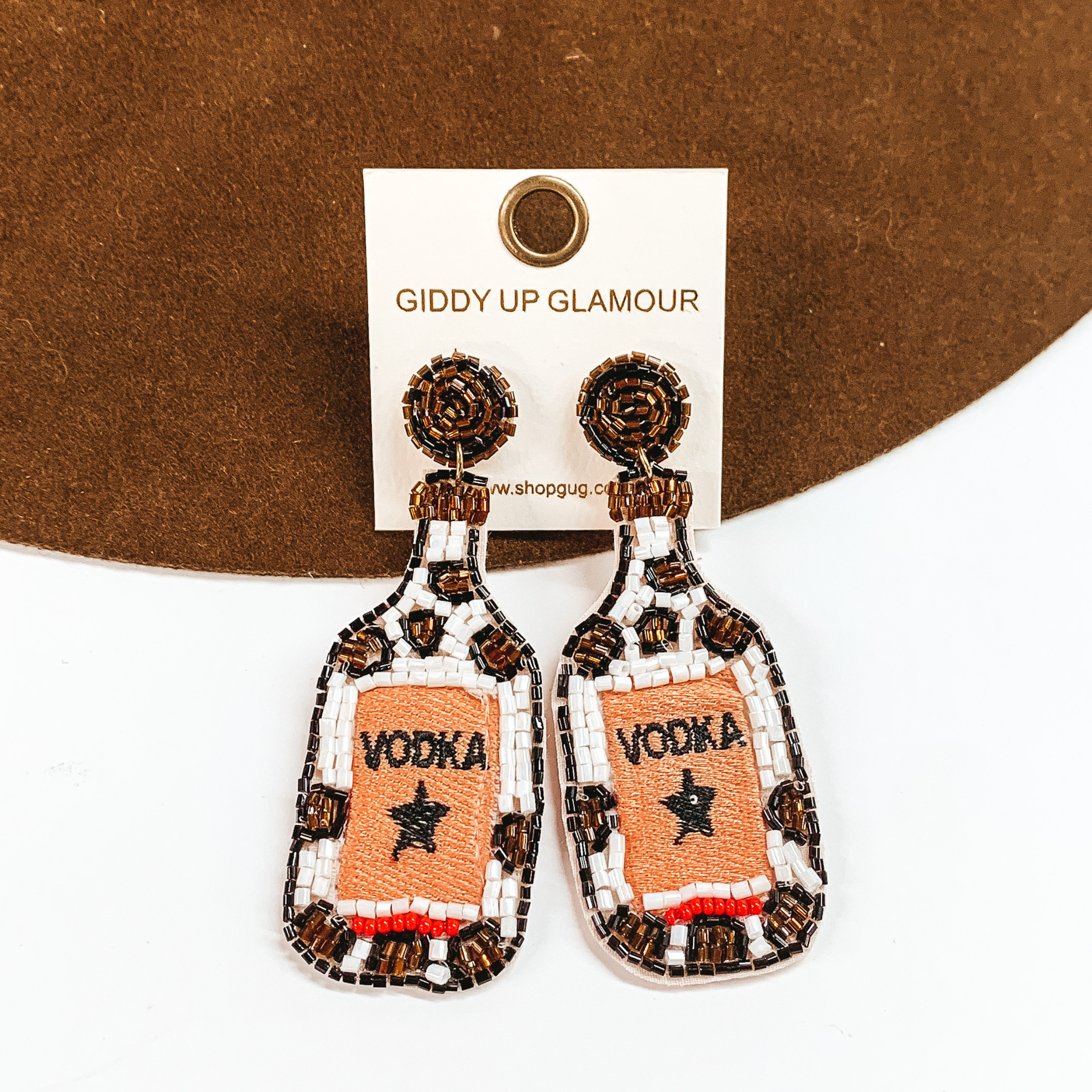 White beaded bottle earrings with brown and black beaded design. These earrings also include black stitching of a star and the word "VODKA". These earrings are pictured on a white and brown background.