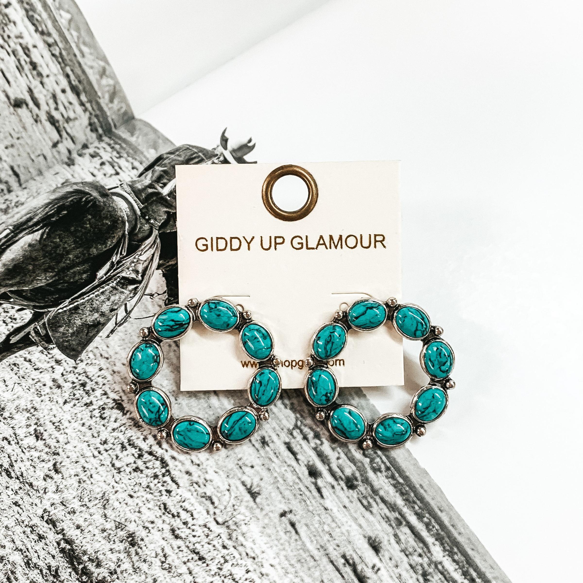 Open circle silver earrings that are outlined in turquoise stones. These earrings are pictured on a black and white picture. 