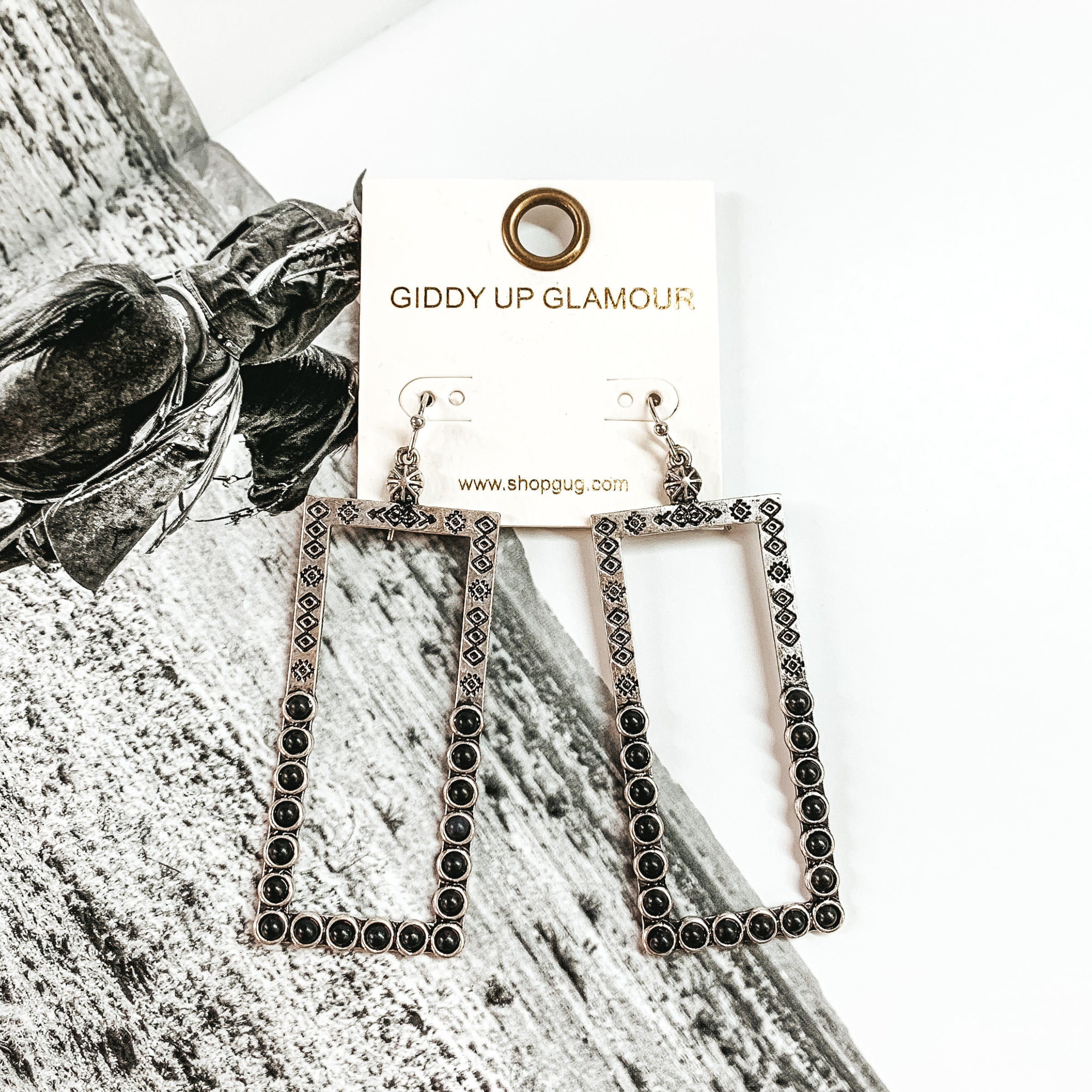 Silver, open rectangle dangle earrings. These earrings include engraving on the top half and black stones on the bottom half. These earrings are pictured on a black and white picture.
