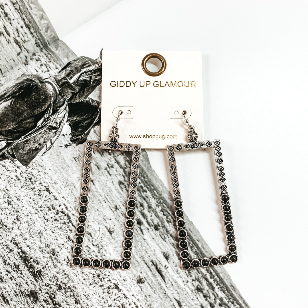 Silver, open rectangle dangle earrings. These earrings include engraving on the top half and black stones on the bottom half. These earrings are pictured on a black and white picture.