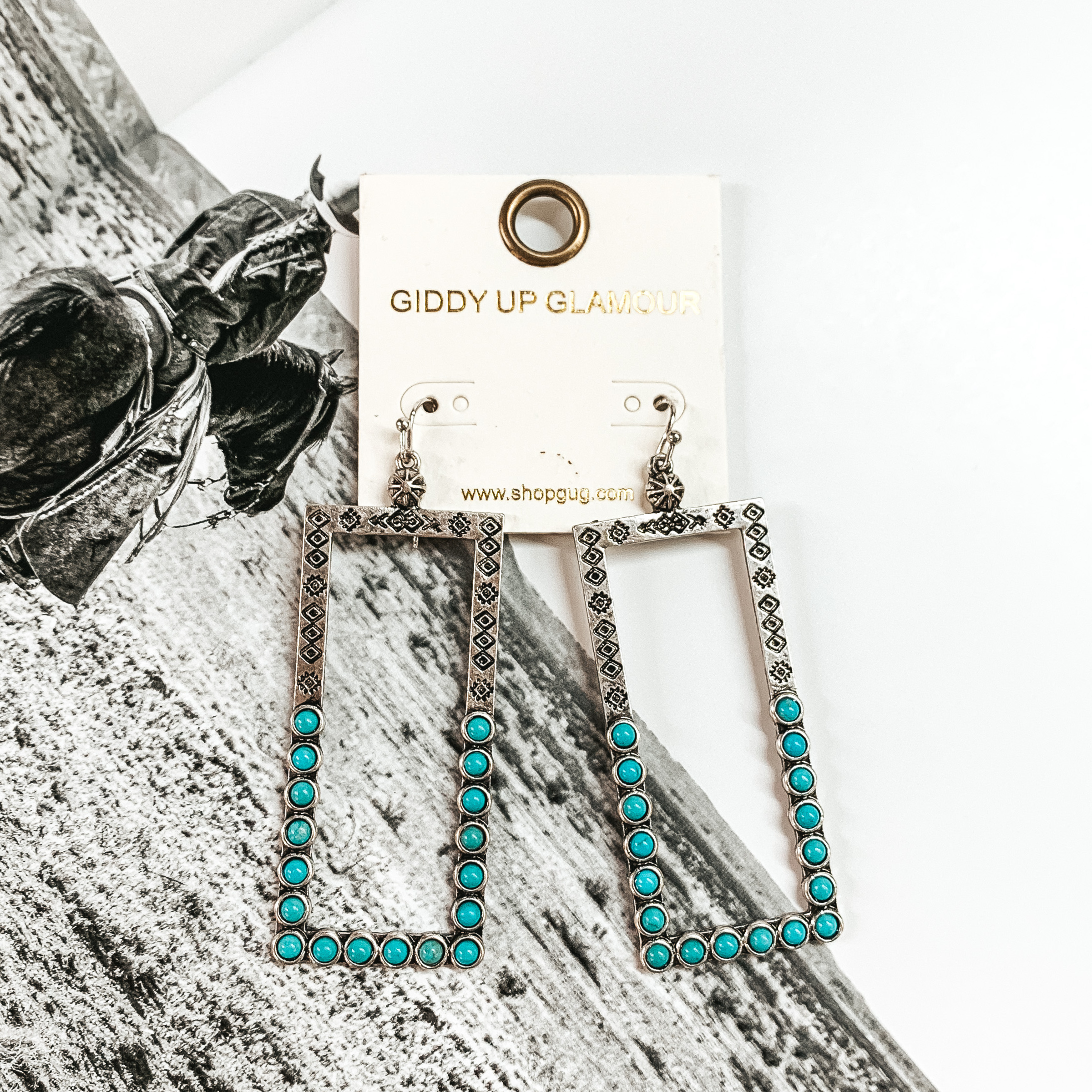 Silver, open rectangle dangle earrings. These earrings include engraving on the top half and turquoise stones on the bottom half. These earrings are pictured on a black and white picture.