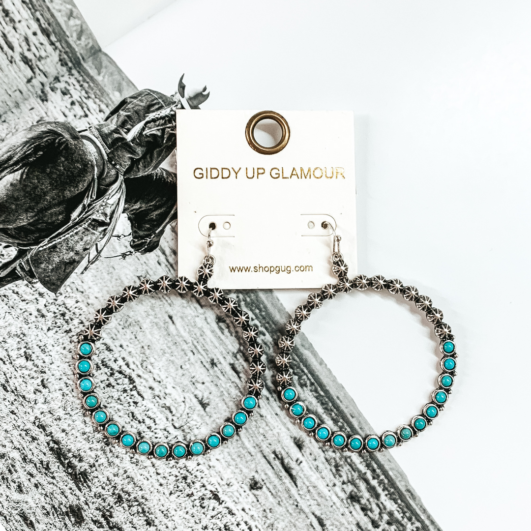 Silver, hoop dangle earrings. These earrings include engraving on the top half and turquoise stones on the bottom half. These earrings are pictured on a black and white picture.