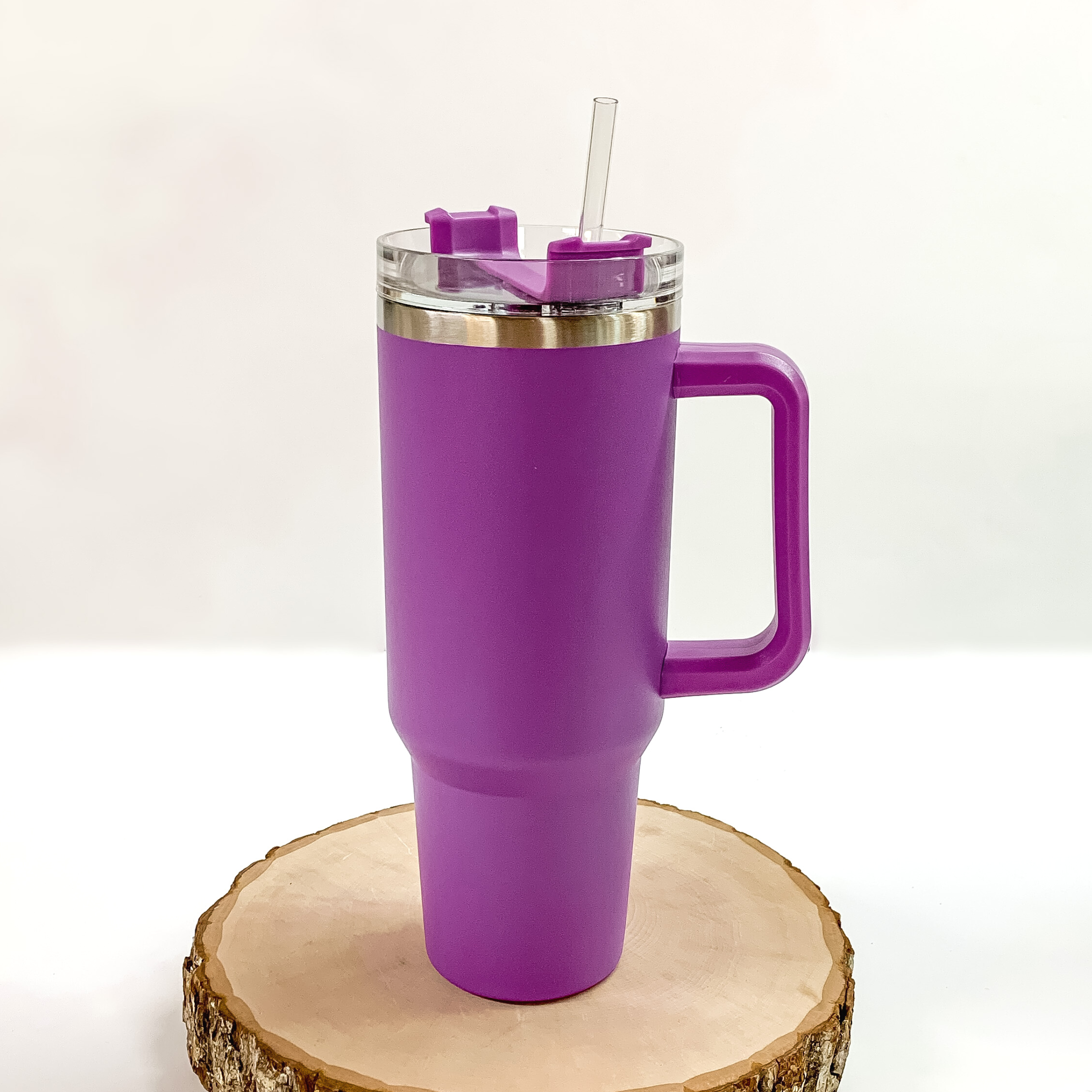 This is a purple tumbler with a purple handle. This tumbler also has a clear lid and straw. This tumbler is pictured standing on a piece of wood on a white background. 