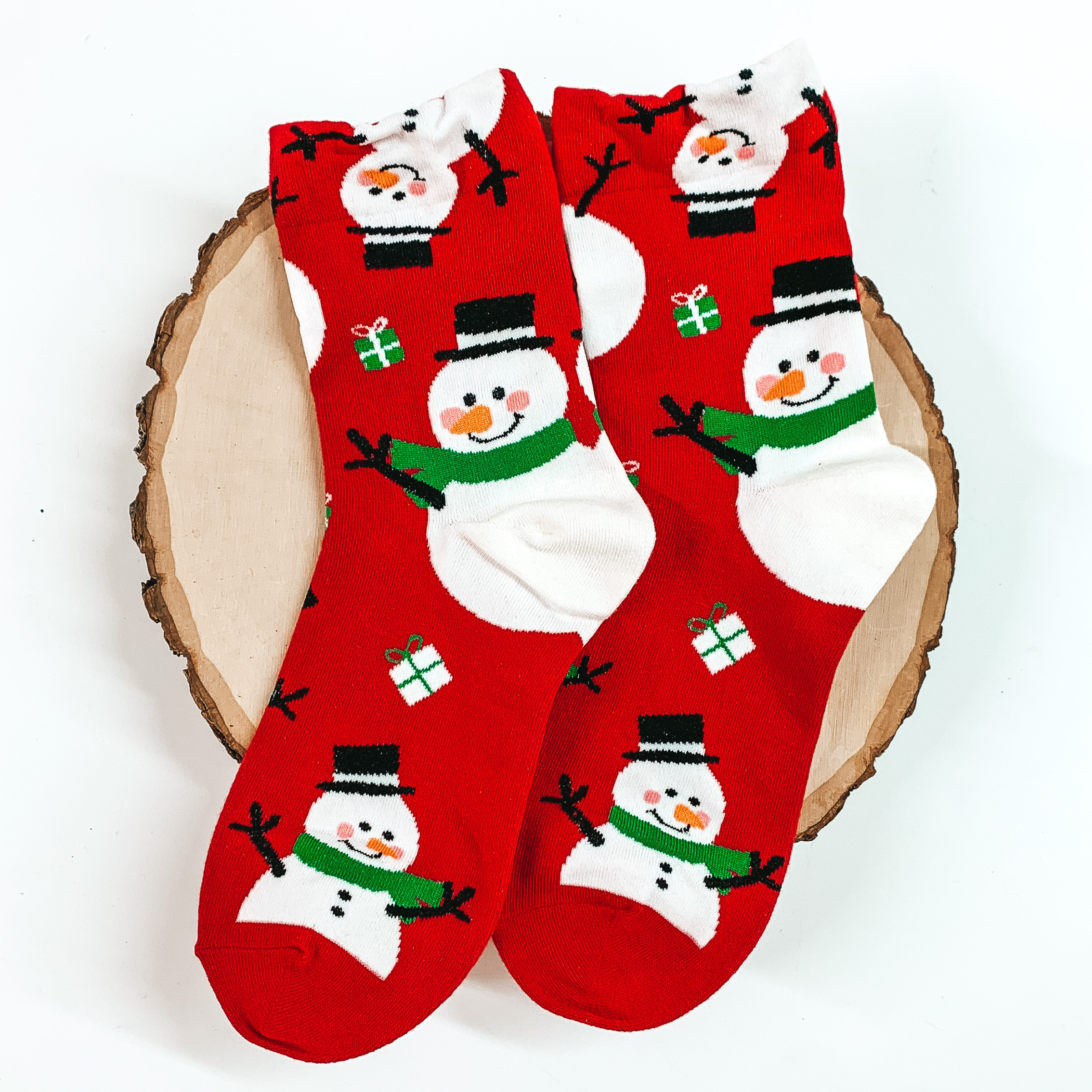 Red ankle socks that have a snowman print. These snowmans include a green scarf and black top hat. These socks are pictured on a piece of wood on a white background. 