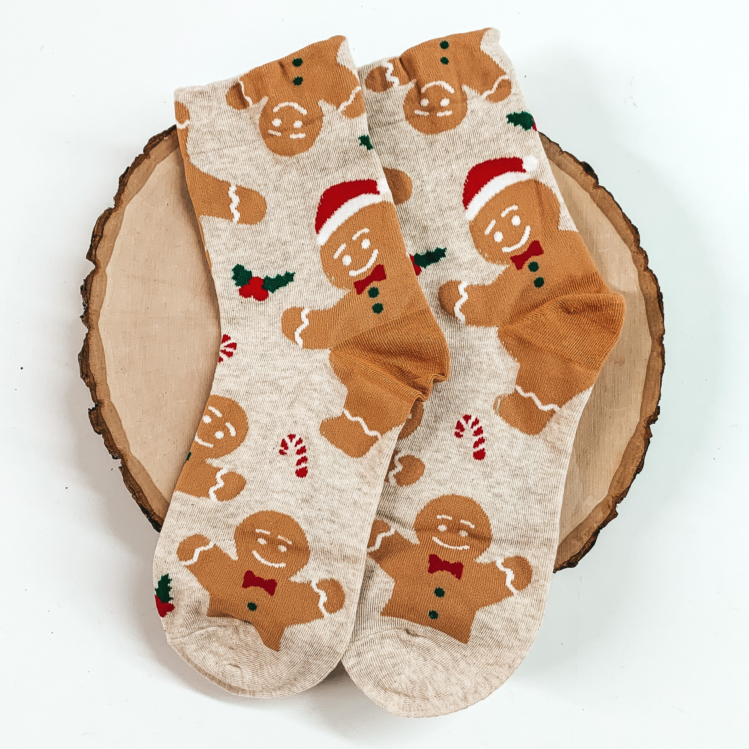 Beige ankle socks with a gingerbread print with candy canes and mistletoe. These socks are pictured on a piece of wood on a white background. 