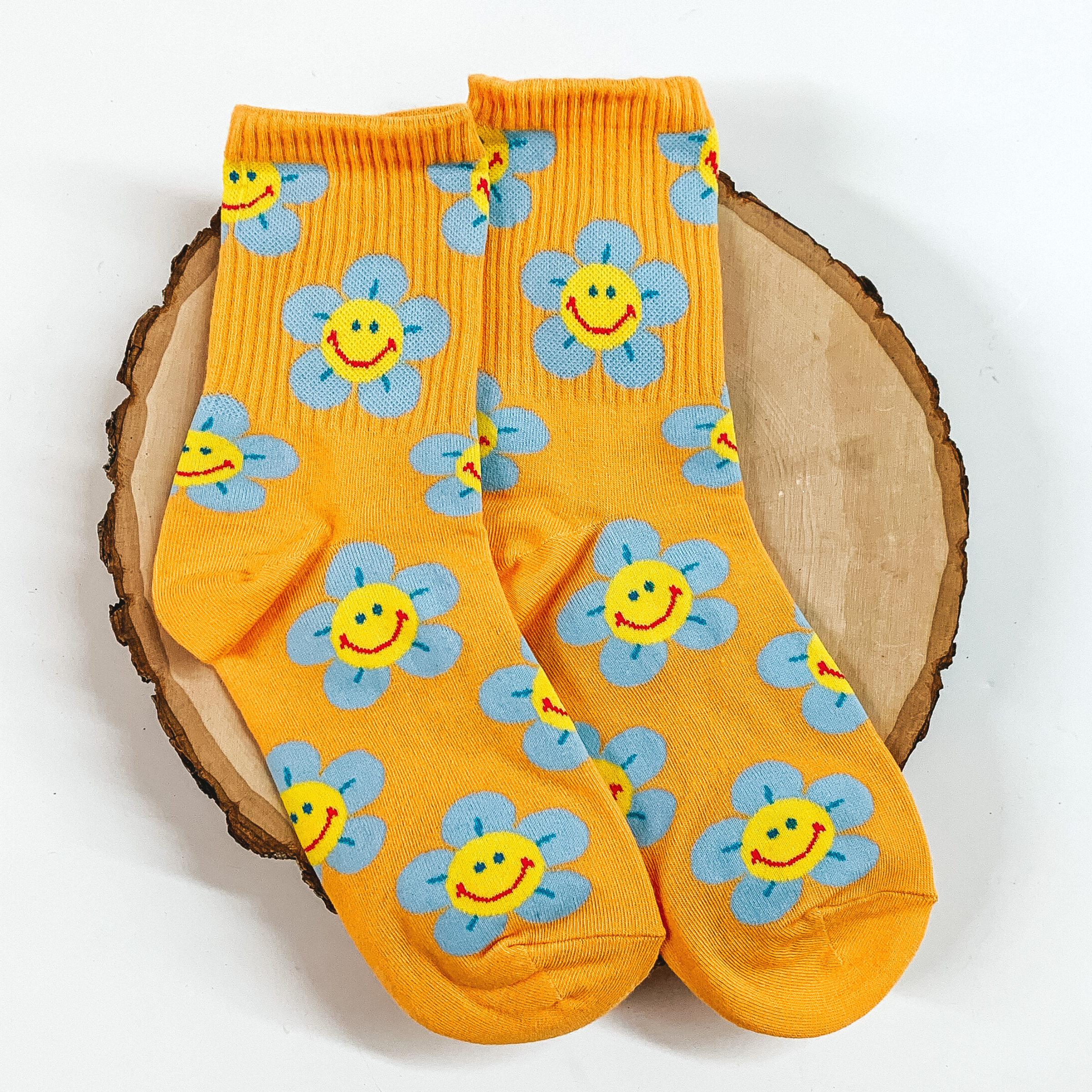 Orange ankle socks with a blue flower print. The flowers have a smiley face in the center. These socks are pictured on a piece of wood on a white background. 